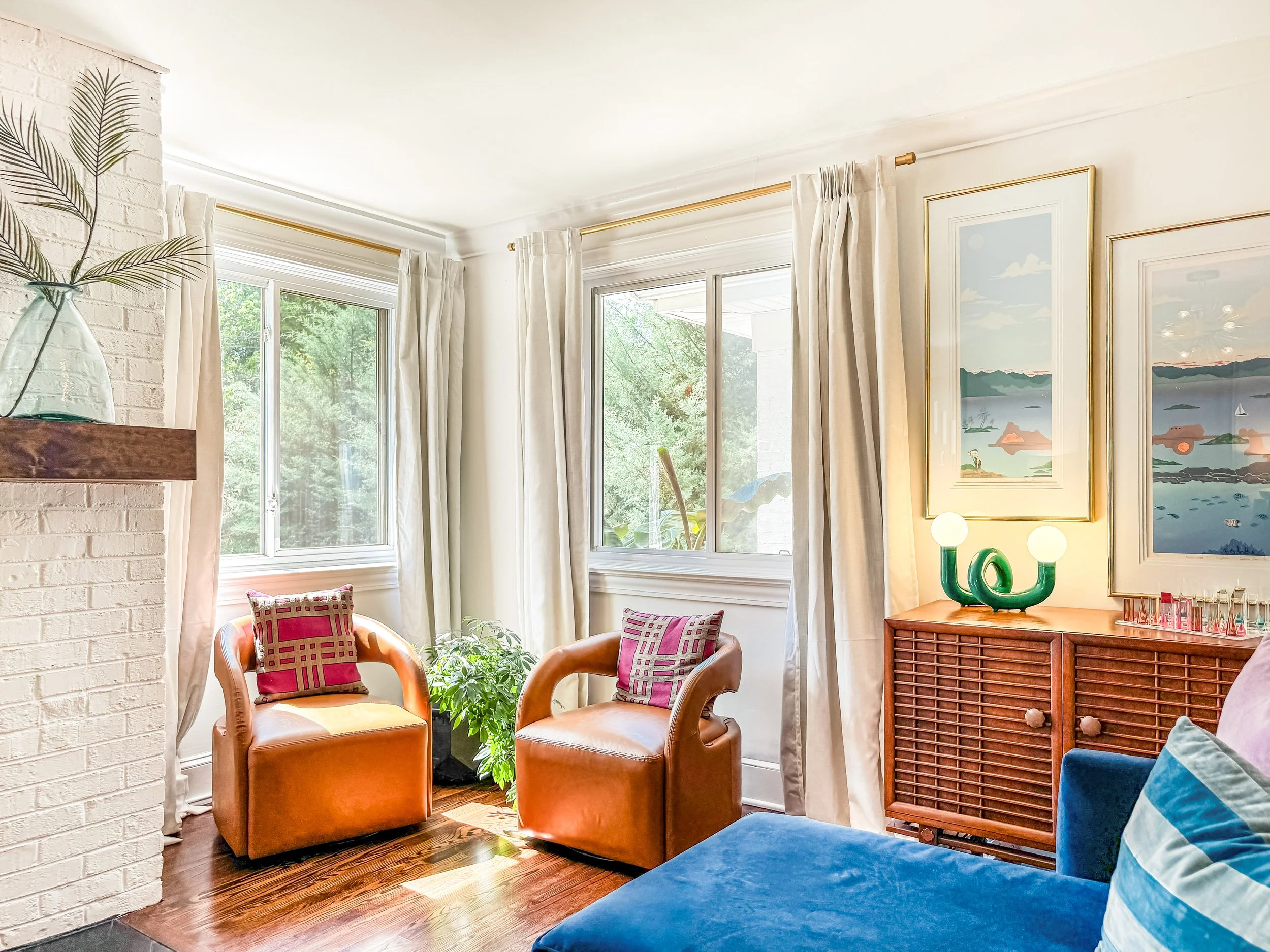 A bright living room with white walls and large windows with white curtains, featuring two tan armchairs with red and purple pillows, a blue sofa with a striped pillow, a wooden sideboard with a green lamp, floral artwork, a potted plant, and a white brick fireplace with a glass vase and plant on the mantel.