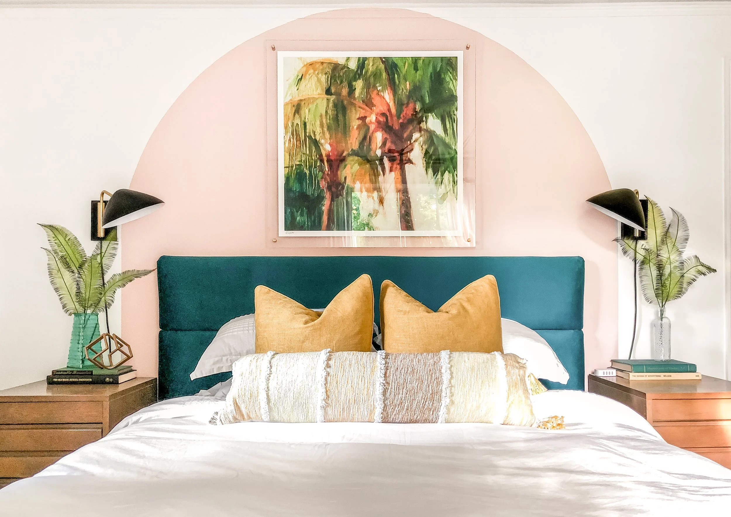 A bedroom with a teal upholstered headboard, large yellow pillows, and a gold and white decorative pillow. There are two wooden nightstands with books, green vases with tropical leaves, and black wall-mounted lamps. A framed painting of palm trees hangs above the bed, and the wall behind has a pink semi-circular accent.