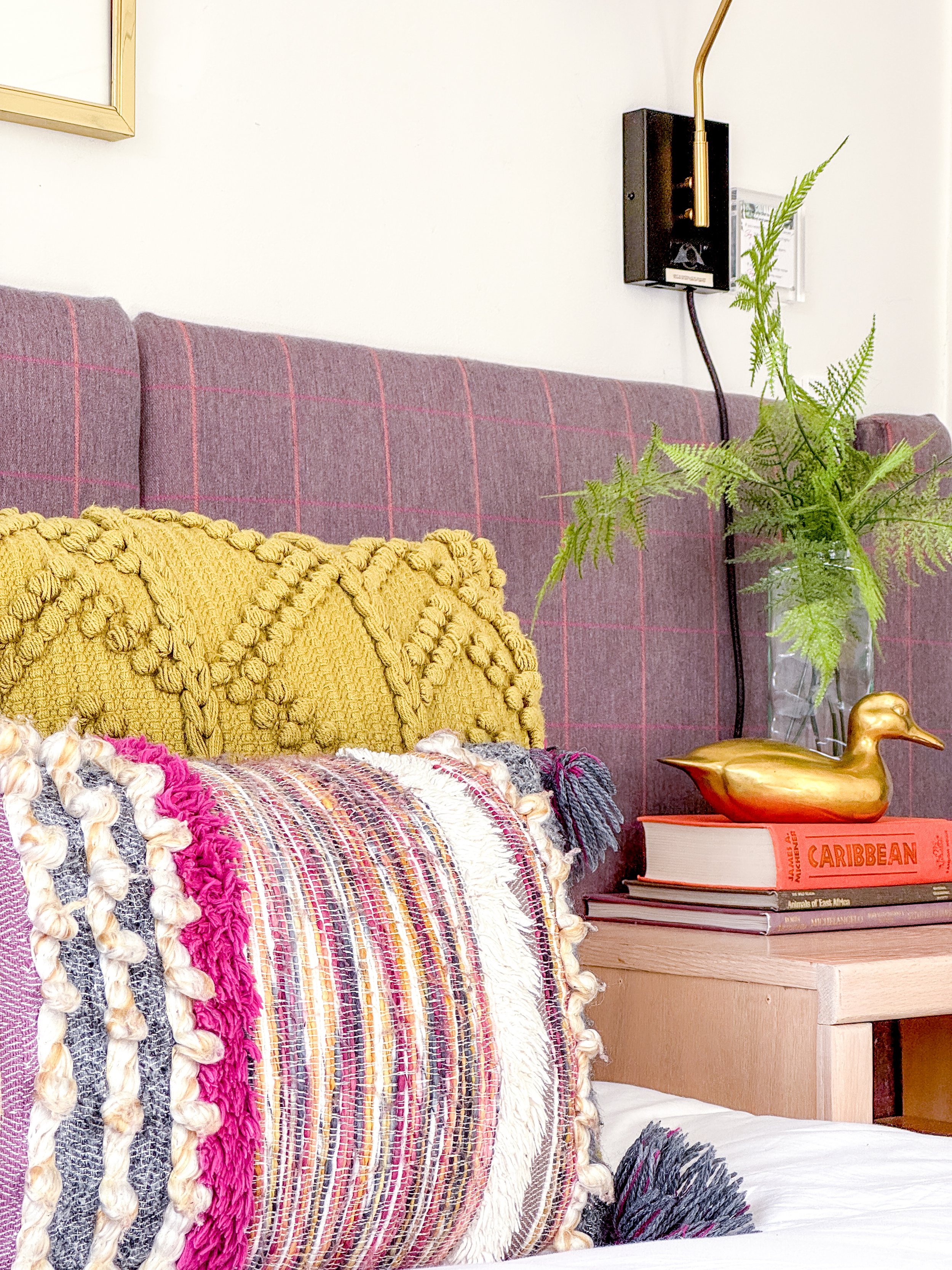 A bedroom corner with a pink upholstered headboard, a textured yellow pillow, a colorful woven throw blanket, a wooden nightstand with a gold duck figurine, a large book titled "Caribbean," a clear vase with green fern leaves, and a wall-mounted black device with a gold lamp above.