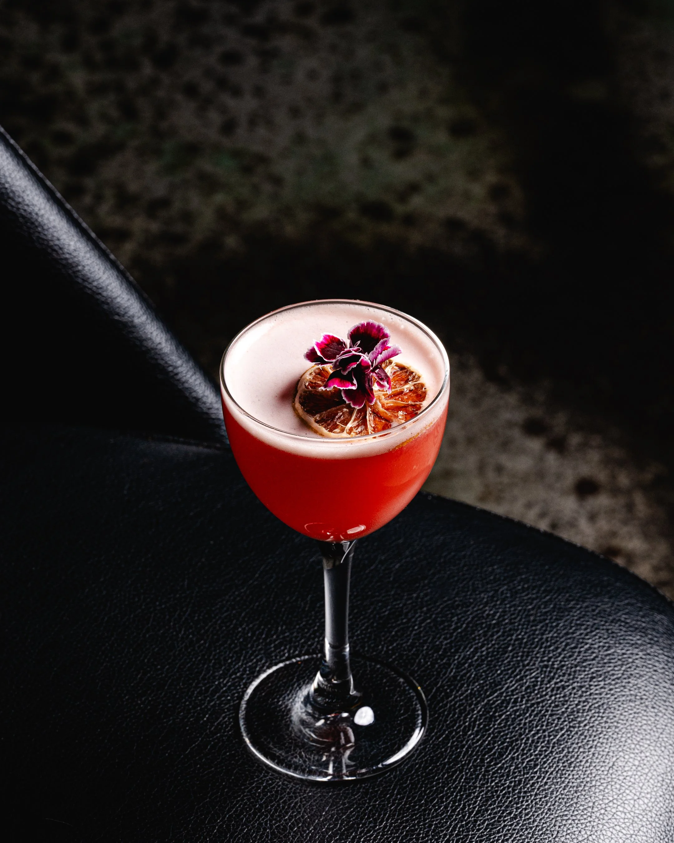 A pink cocktail garnished with dried citrus slices and purple flowers in a wine glass.