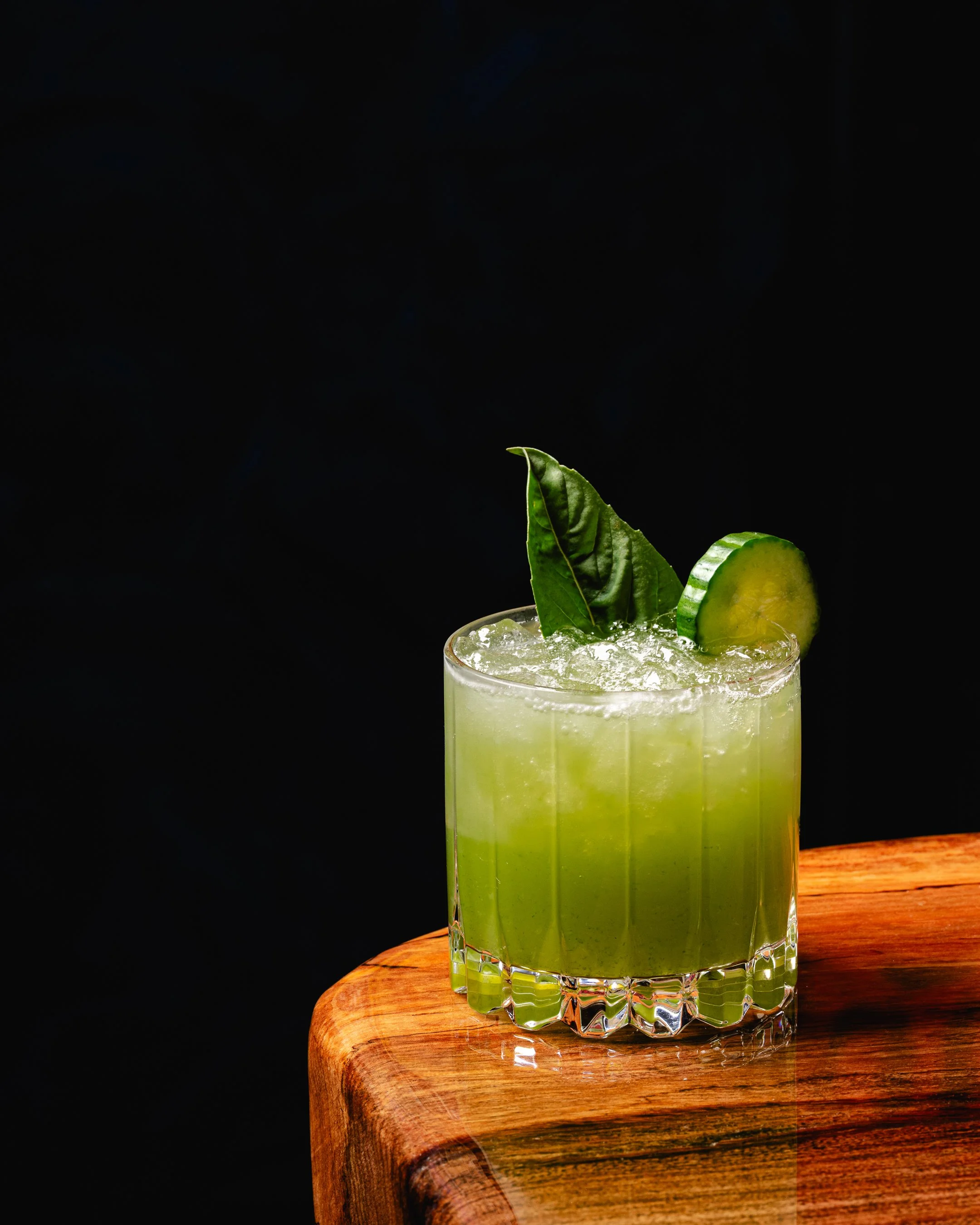 A green cocktail garnished with cucumber slice and basil leaves on a wooden table with a black background.