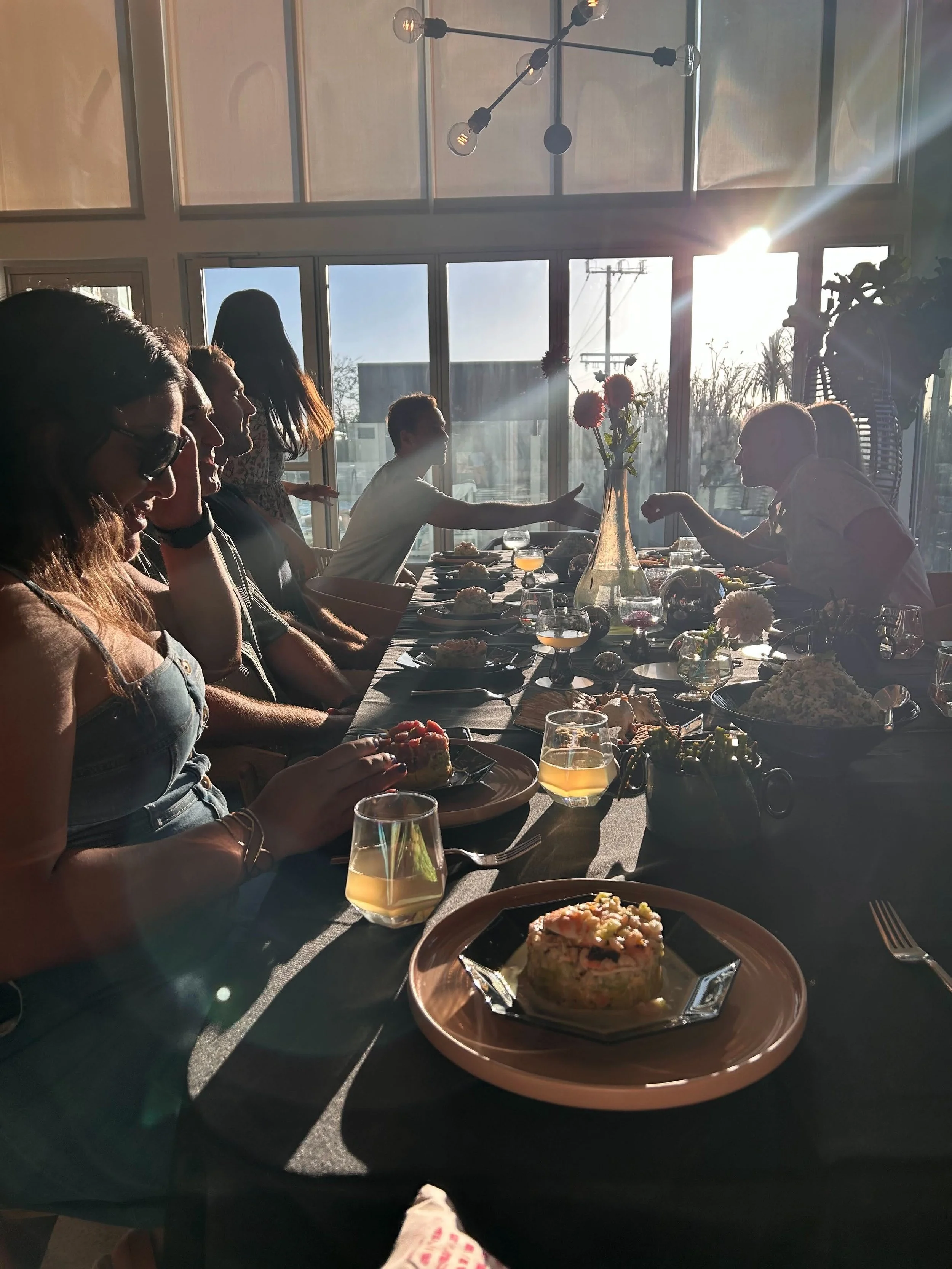 People sitting around a dining table enjoying a meal, with sunlight shining through large windows in the background.