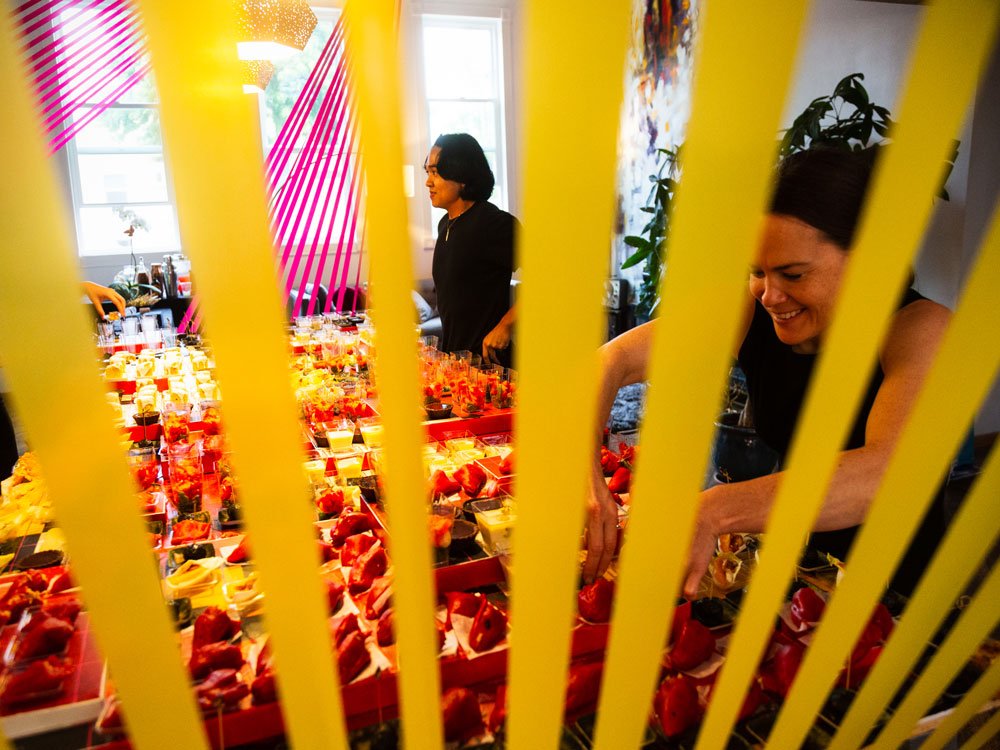 Two women in black shirts preparing and arranging small pre-packaged food items in a room decorated with yellow and pink accents.