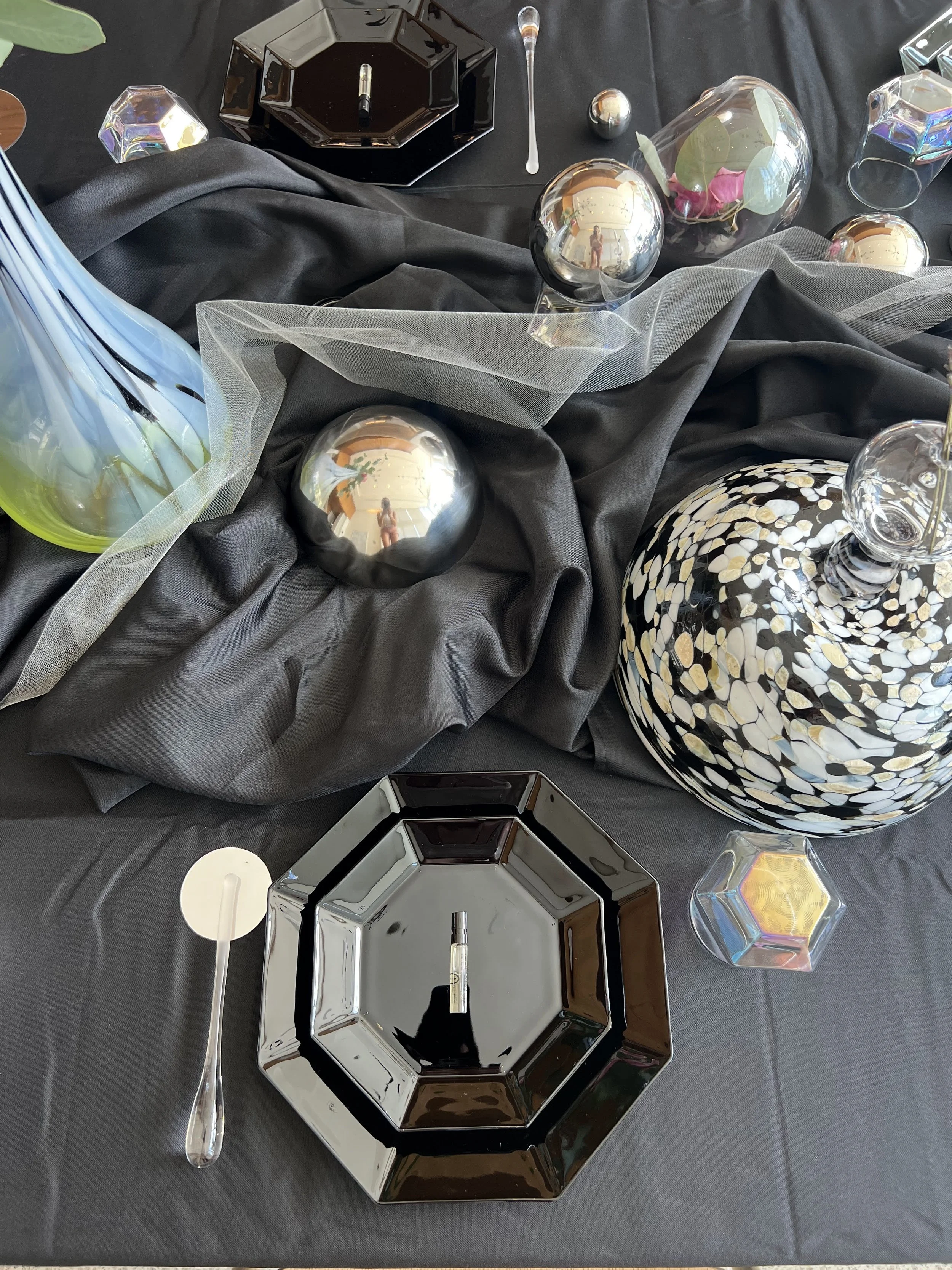 Decorative table setting with black and white ceramic plates, reflective silver and holographic balls, glass vases with floral arrangements, and various geometric glass containers, all on a black cloth with draped fabric.
