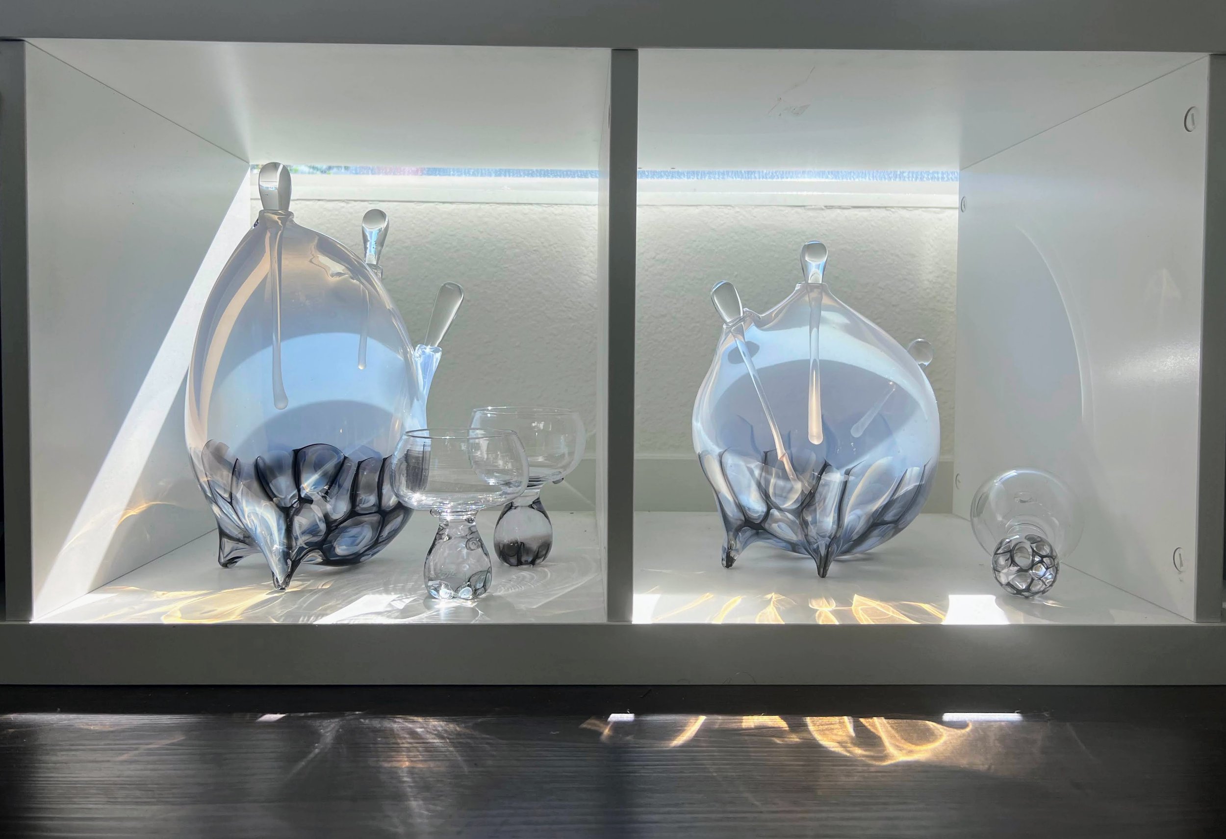 Display case with two large, artistic glass vases and several small glass objects, all made of clear and smoky glass, with reflections and light patterns.