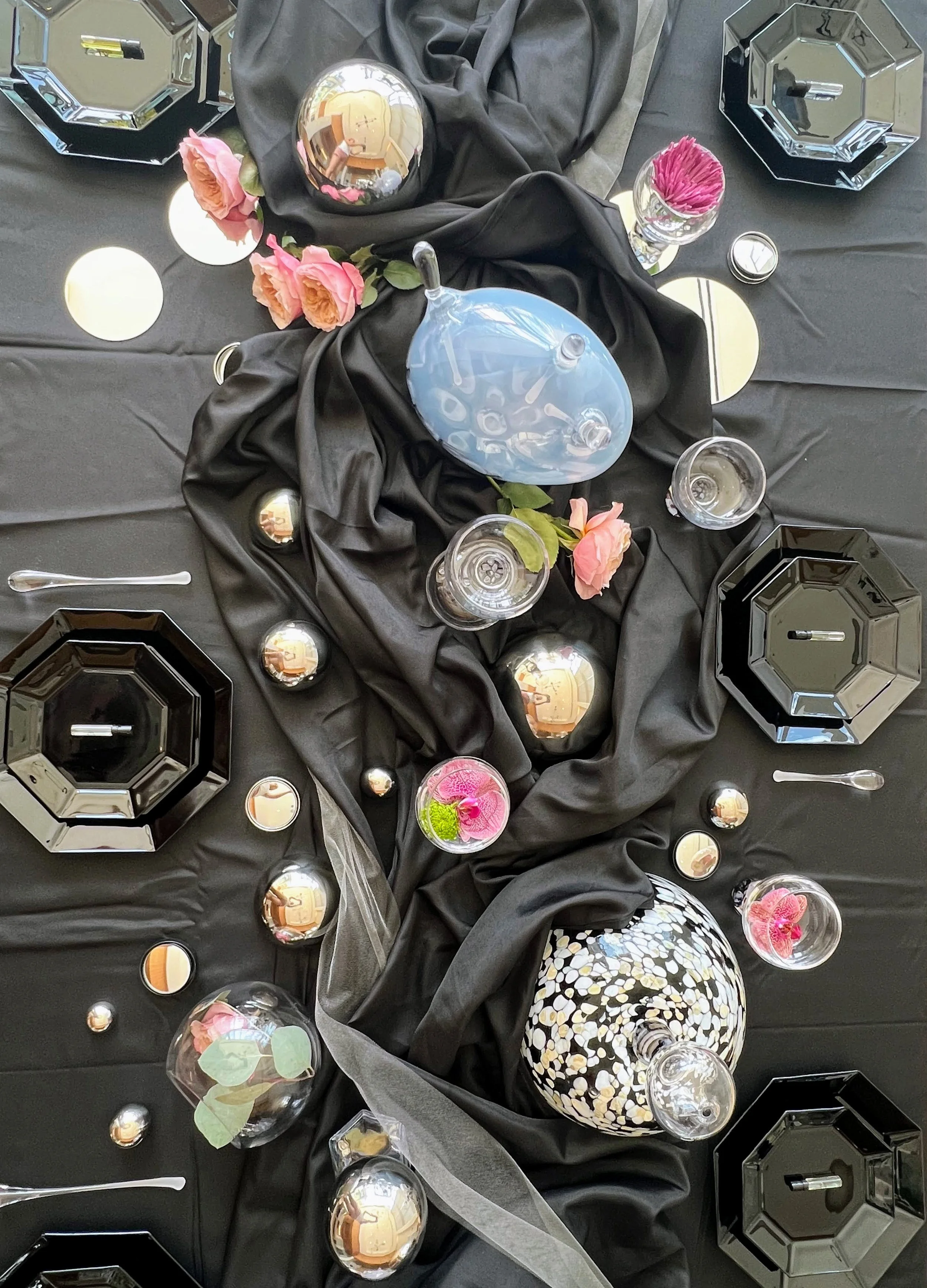 A decorative table centerpiece with black fabric and metallic and glass accents, including mirrored spheres, glass bowls of flowers, and black geometric containers.