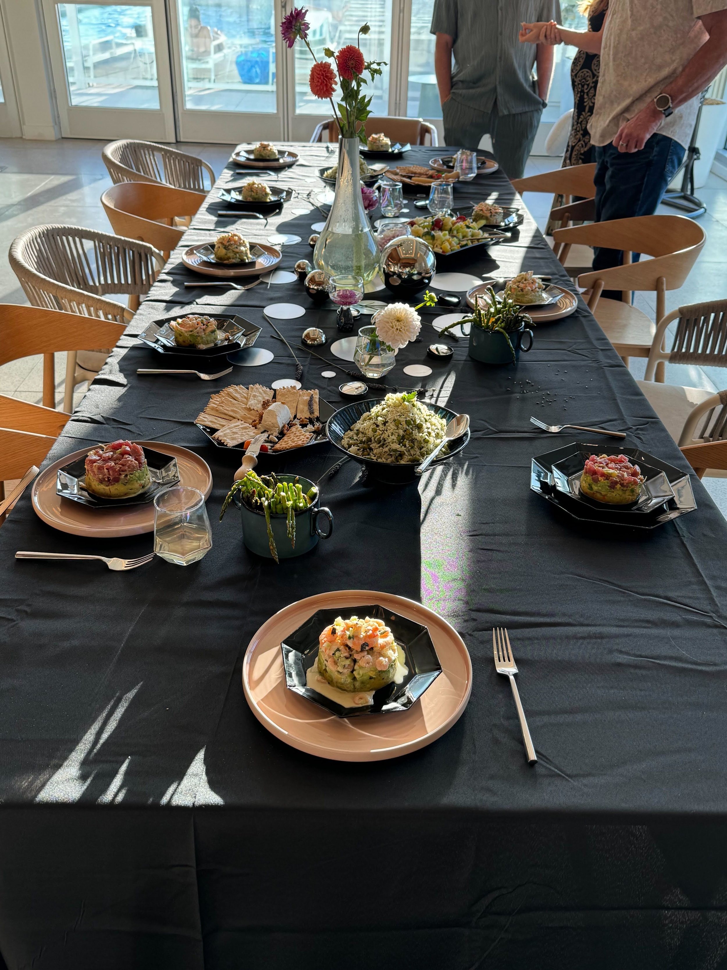 A dining table set for a gathering, with plates of food, flowers in a vase, and scattered decorative objects, in a room with large windows and sunlight.