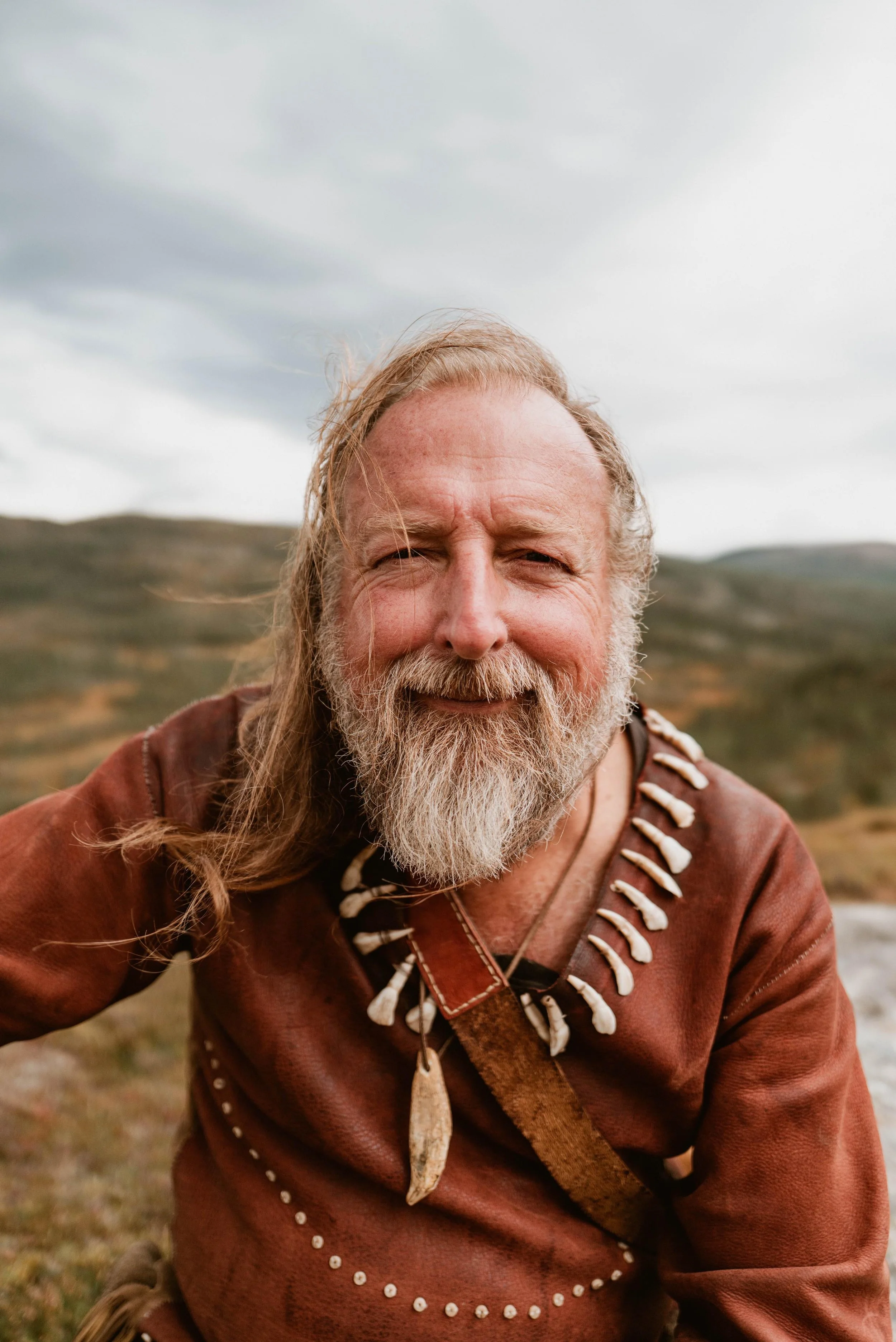Man wearing traditional leather clothing with bone decorations, outdoors in a natural setting.