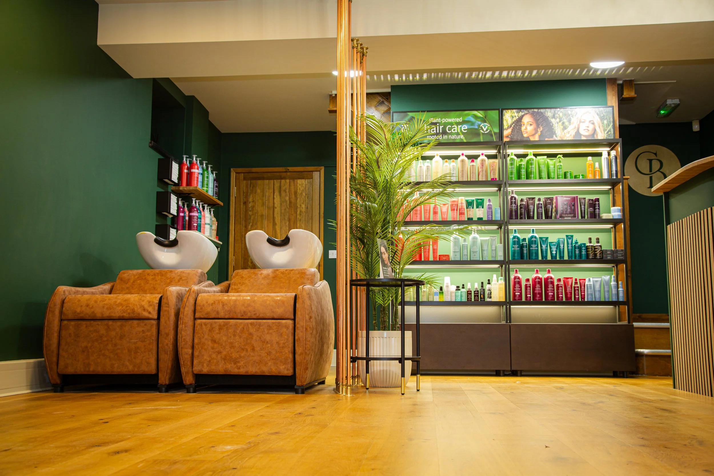 Interior of a hair salon with two brown armchairs under hair washing sinks, a wooden door, and shelves stocked with colorful hair care products, a potted plant in the center, and a wooden floor.