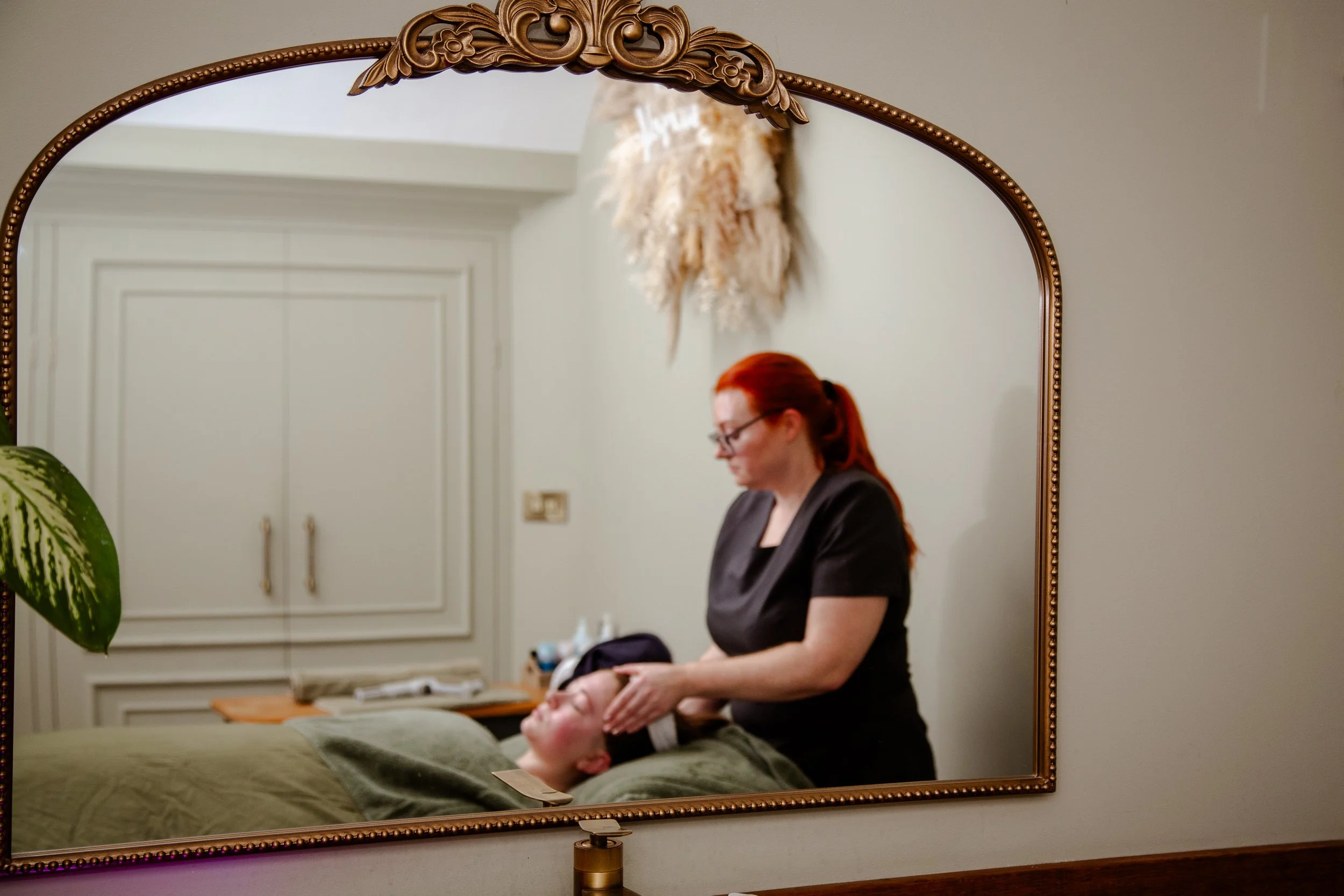 A woman with red hair and glasses giving a facial treatment to a client lying on a bed, reflected in an ornate framed mirror.