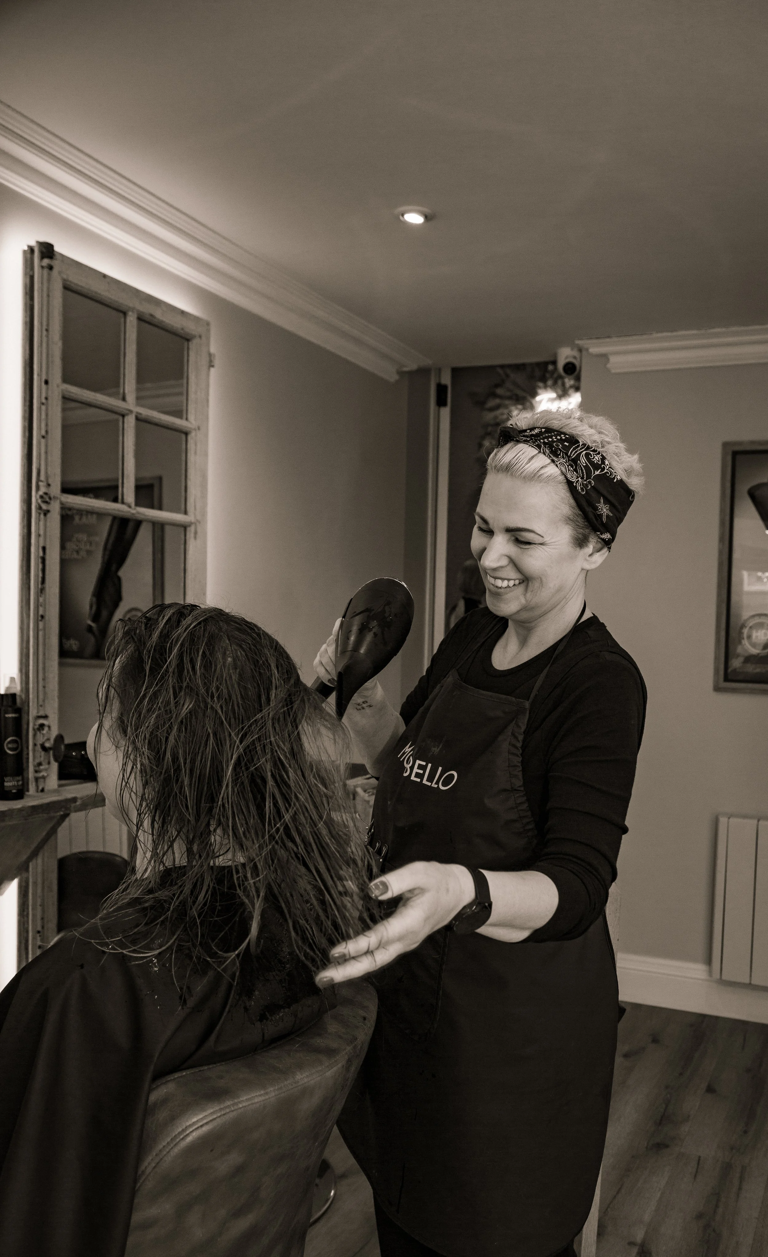 A hairstylist with a bandana styling a woman's wet hair in a salon with a mirror and window in the background.