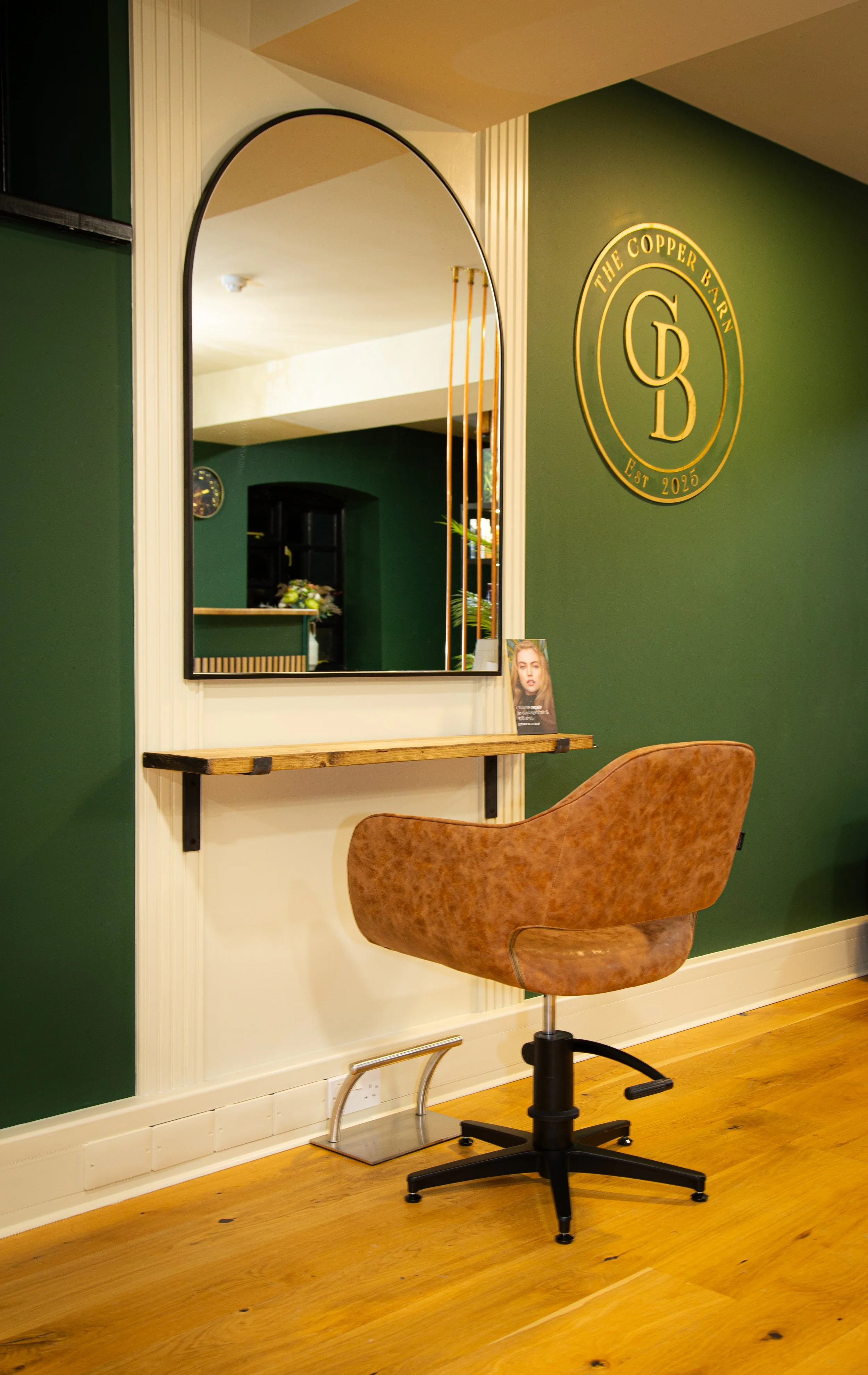 Interior of a room with a green wall, a mirror, a wooden shelf, and a brown swivel chair on a wooden floor. There is a logo on the wall that reads 'The Copper Bank Est 2025'.