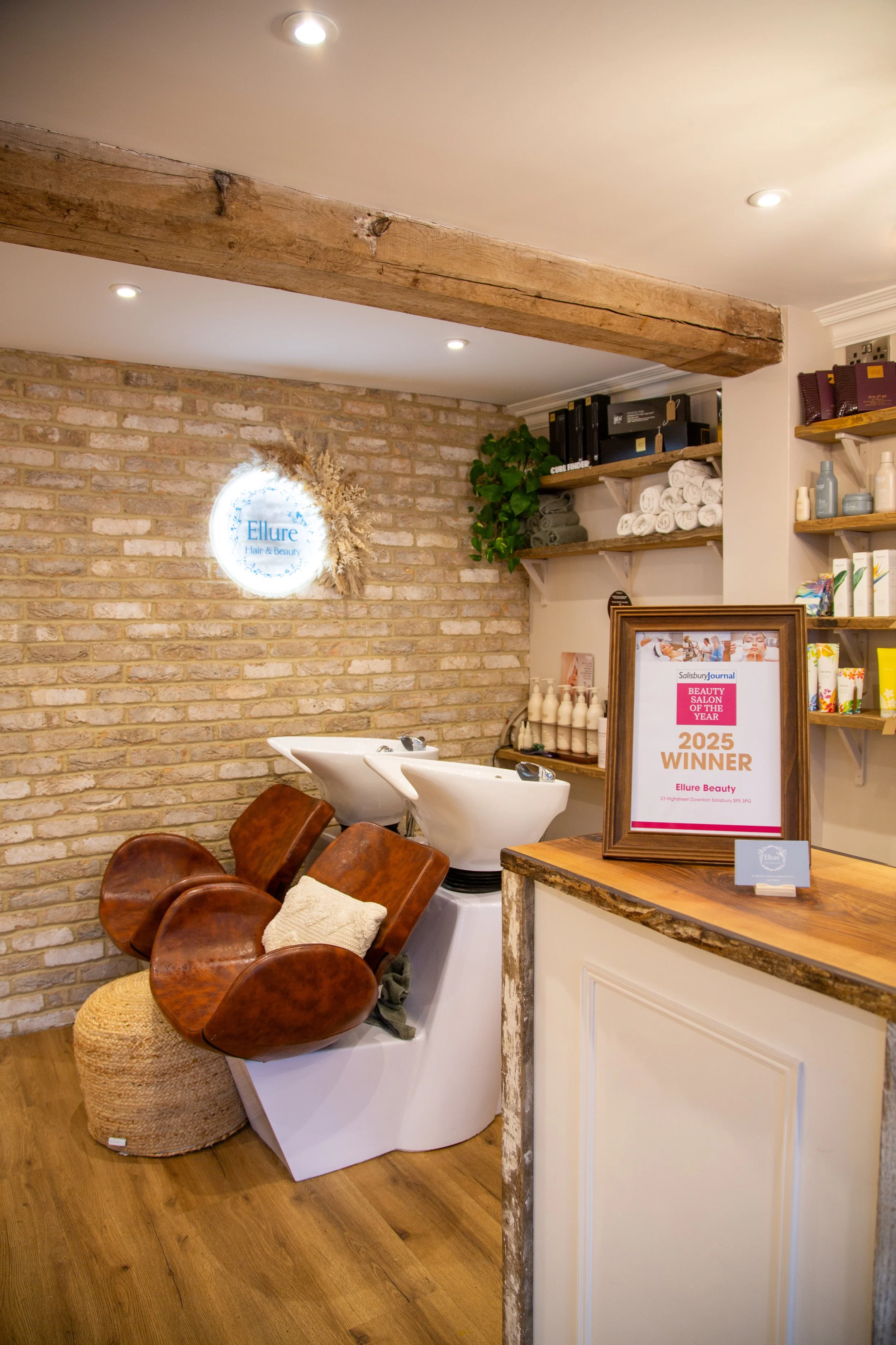 Interior of a hair and beauty salon with shampoo stations, a brick wall, a framed award plaque, and shelves with beauty products and towels.