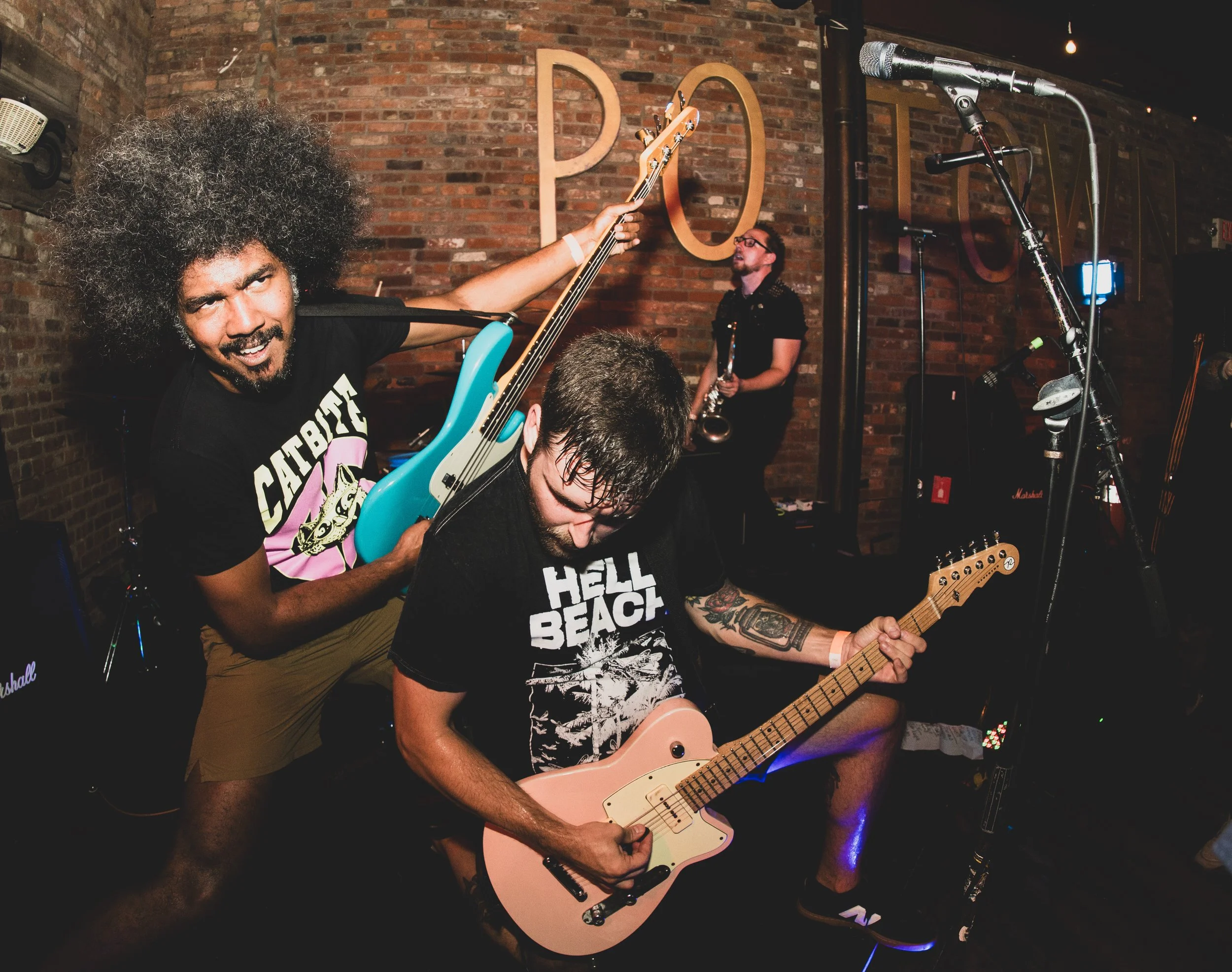Two musicians performing passionately on stage in a brick-walled venue. One with a large afro and wearing a black 'CATABBA' t-shirt is holding a blue bass guitar, and the other with tattoos and wet hair is playing a pink electric guitar. A third pers