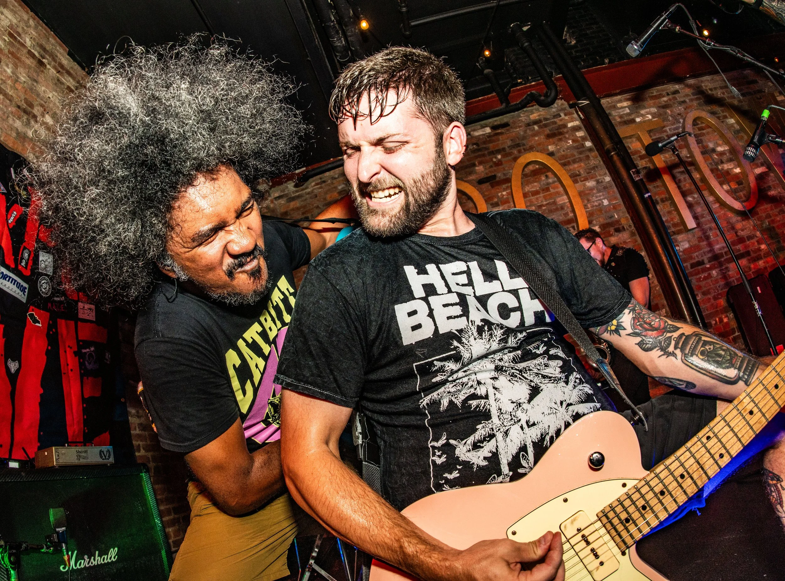 Two musicians performing passionately on stage, one playing an electric guitar and the other singing, with a brick wall background and stage equipment around.