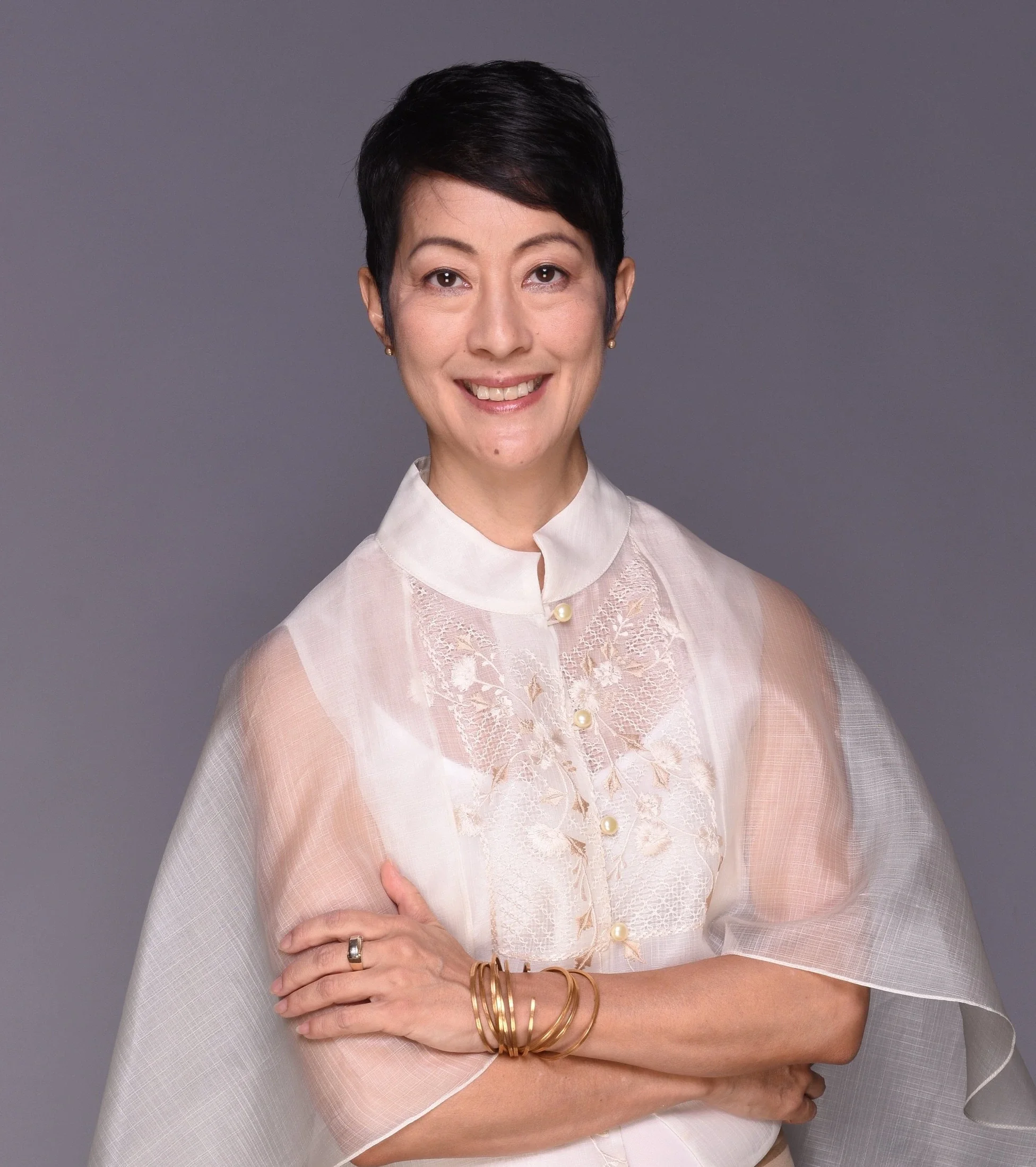 A Japanese-Canadian woman with short black hair smiling, wearing a dressy blouse, against a gray background.