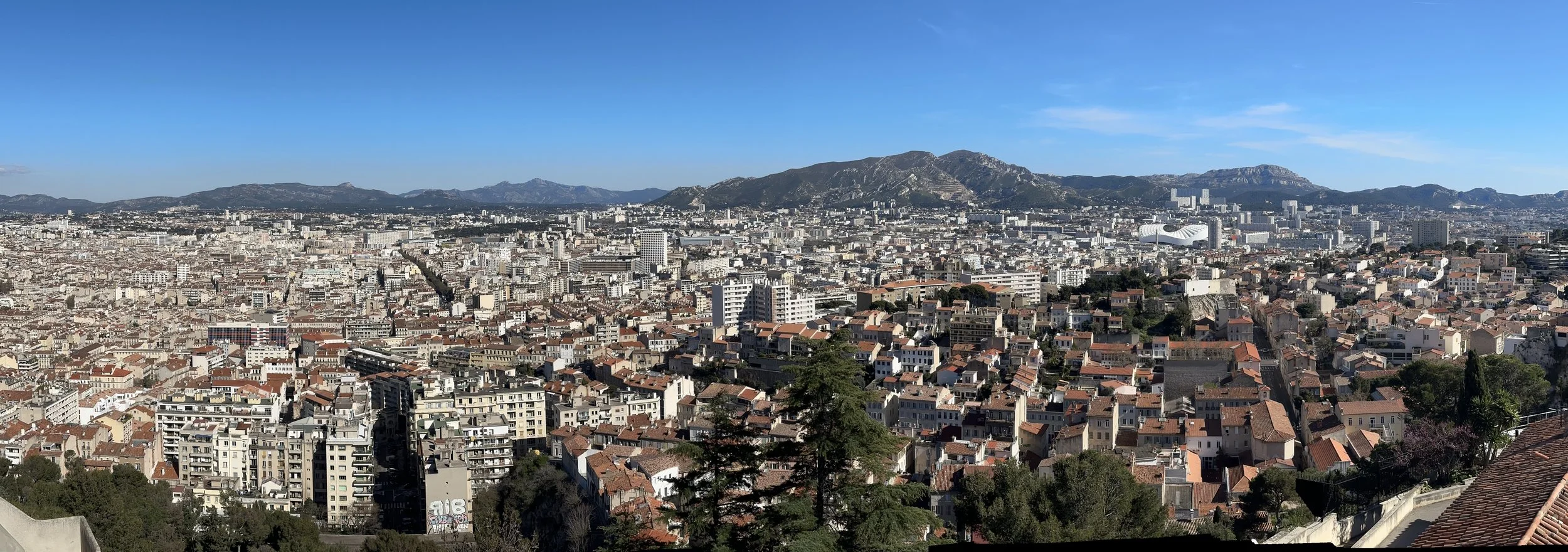 78 - A nice panorama of Marseille, showing the surrounding hills and the "Stade Vélodrome" -- the City's soccer stadium.  The stadium is the large circular white structure at the right of the photo.  The stadium has been home to the Olympique de Mars