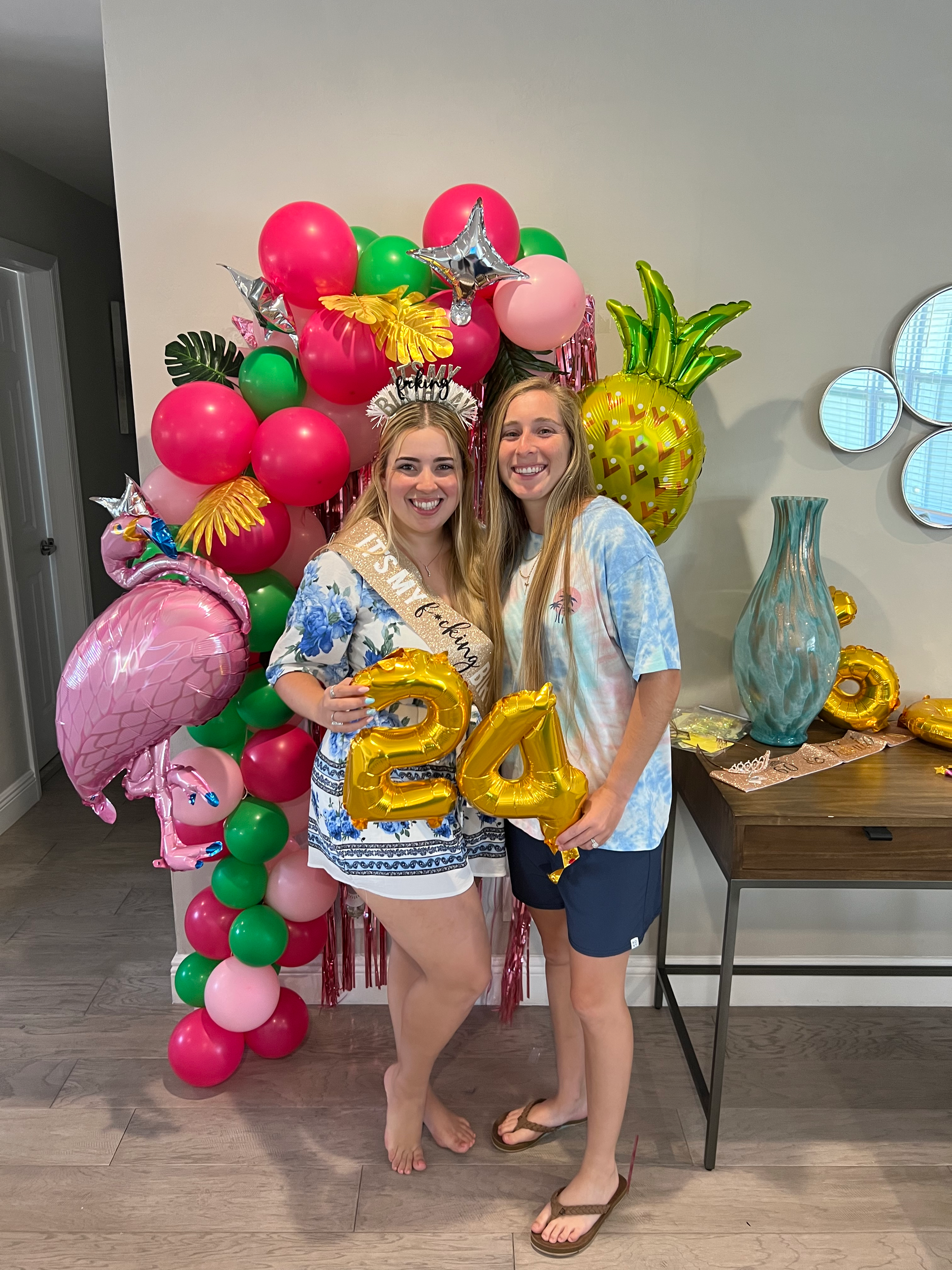 Two women celebrating with colorful balloons, including a pineapple-shaped balloon, wearing casual summer clothes, with one woman holding gold balloons spelling '20', in front of a balloon arrangement with pink, green, and gold decorations, and small