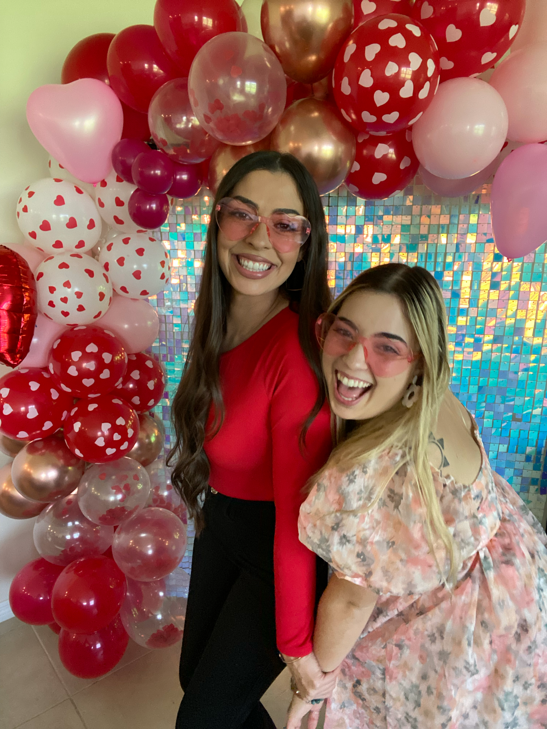 Two women wearing pink-tinted glasses, smiling, and posing in front of a colorful, iridescent mosaic backdrop with pink, red, white, and gold balloons with heart patterns.