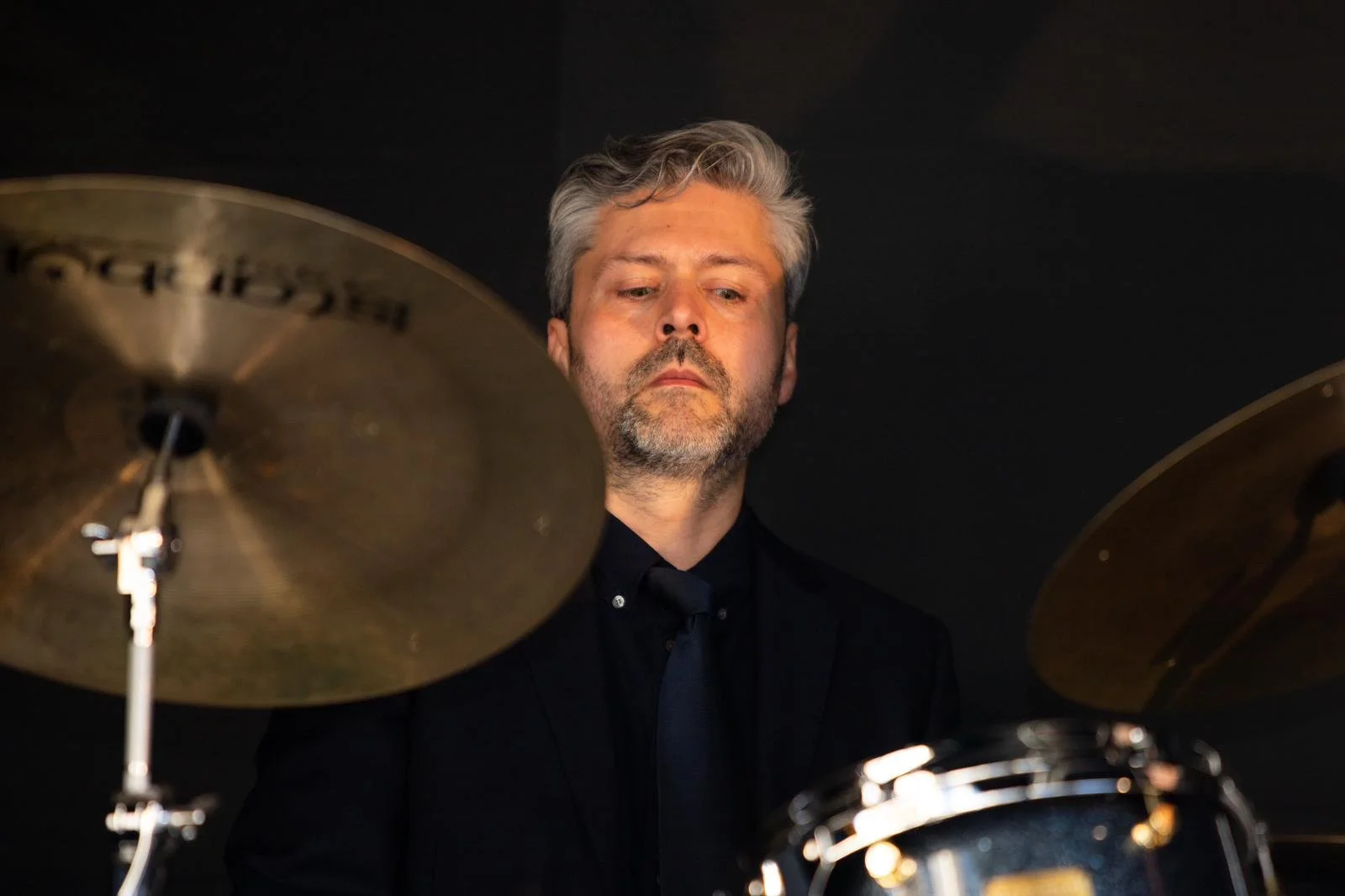 A man with gray hair and a beard playing drums, focusing on his face and drum set against a dark background.