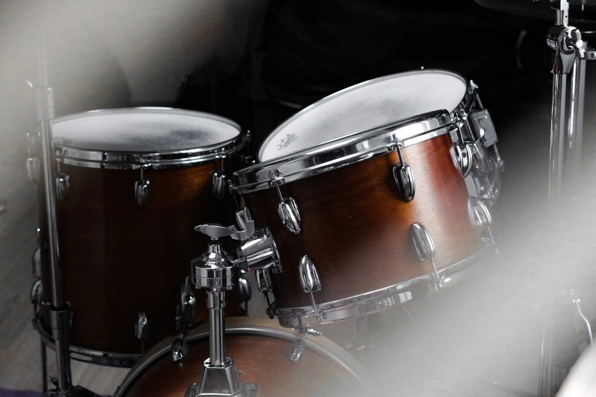 A brown drum set with chrome hardware and white drumheads, placed in a dimly lit room.