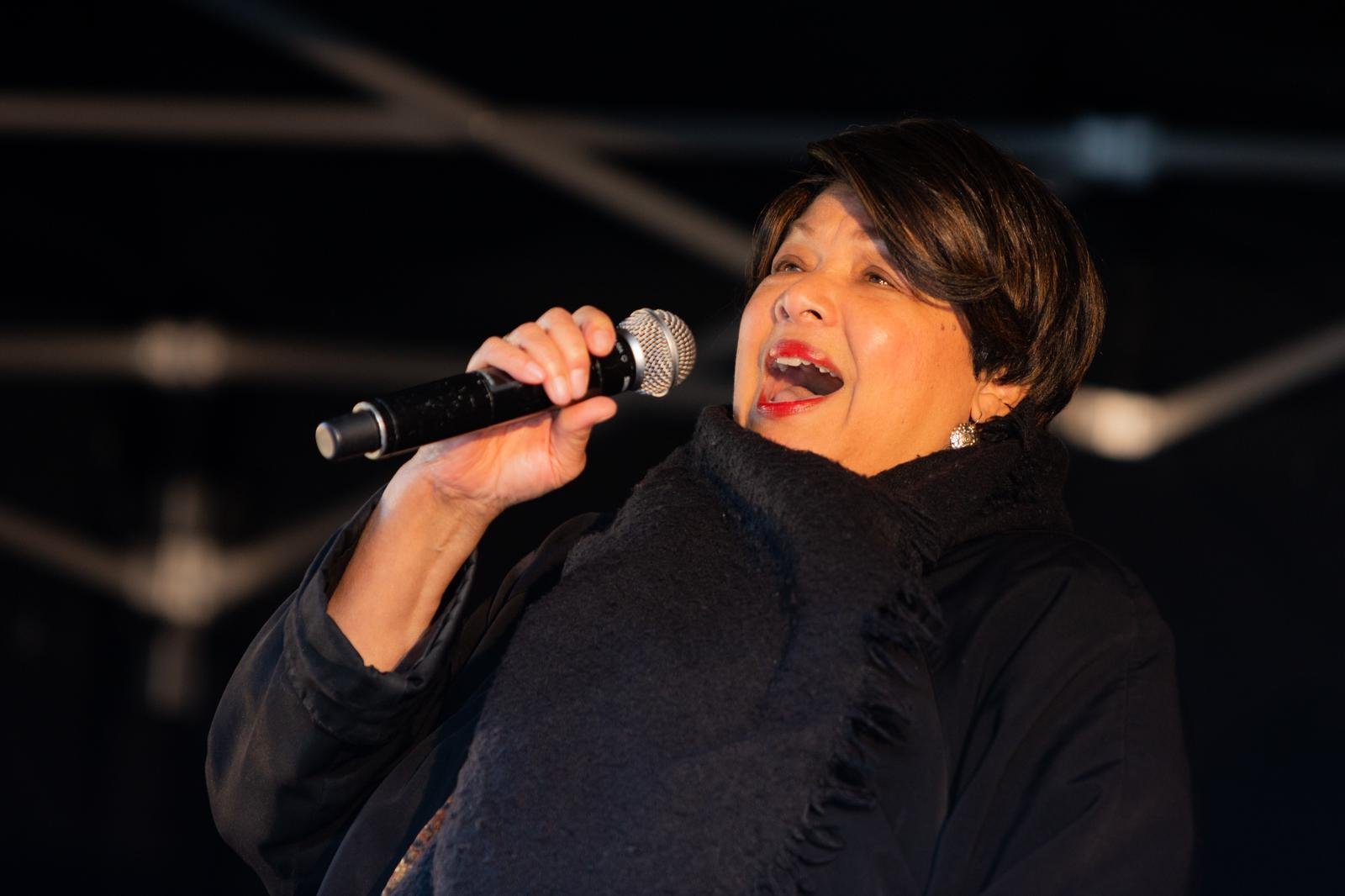 A woman singing into a microphone on stage, wearing a black jacket and having short dark hair.