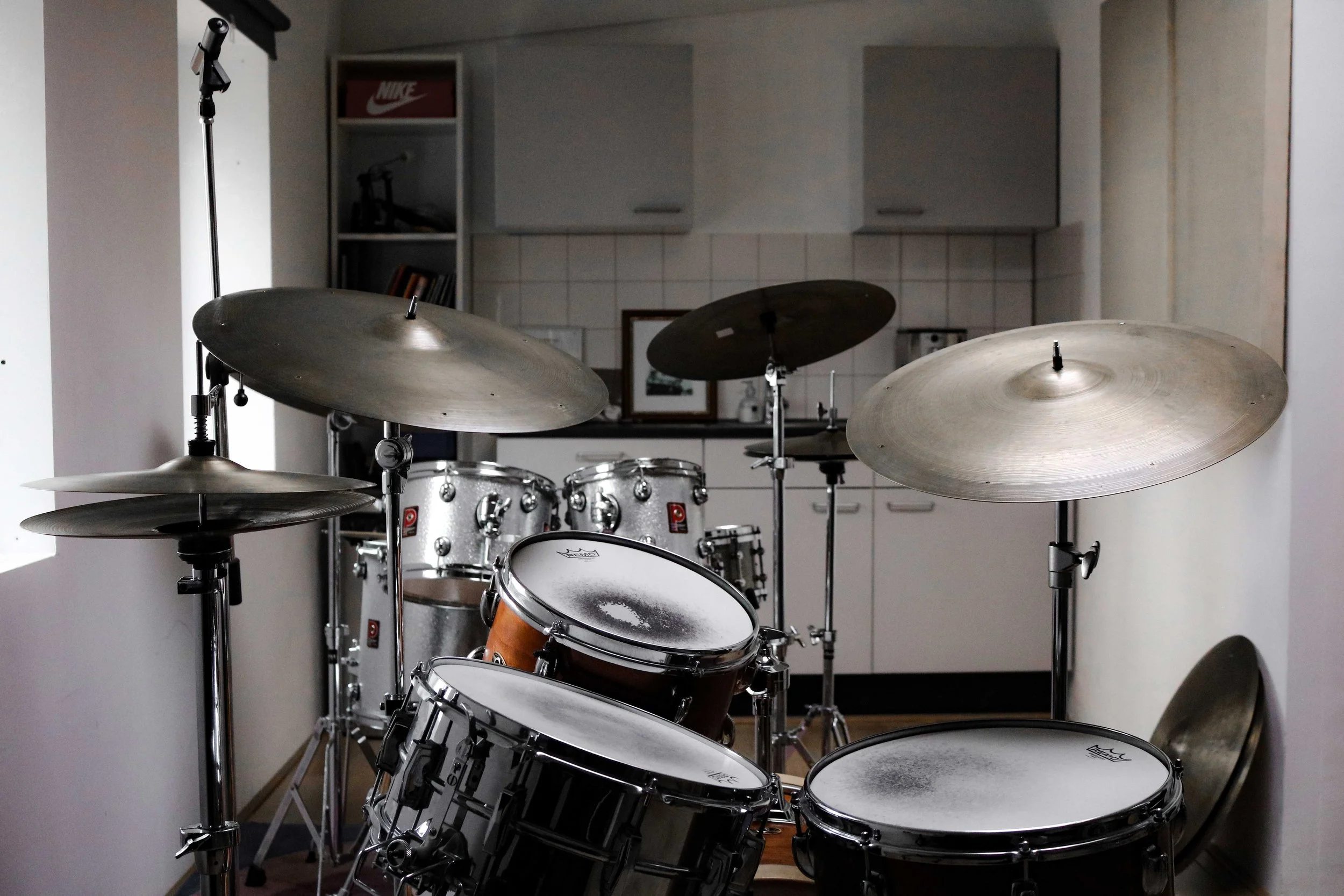 A drum kit with various cymbals and drums set up in a room, with a kitchen area in the background.