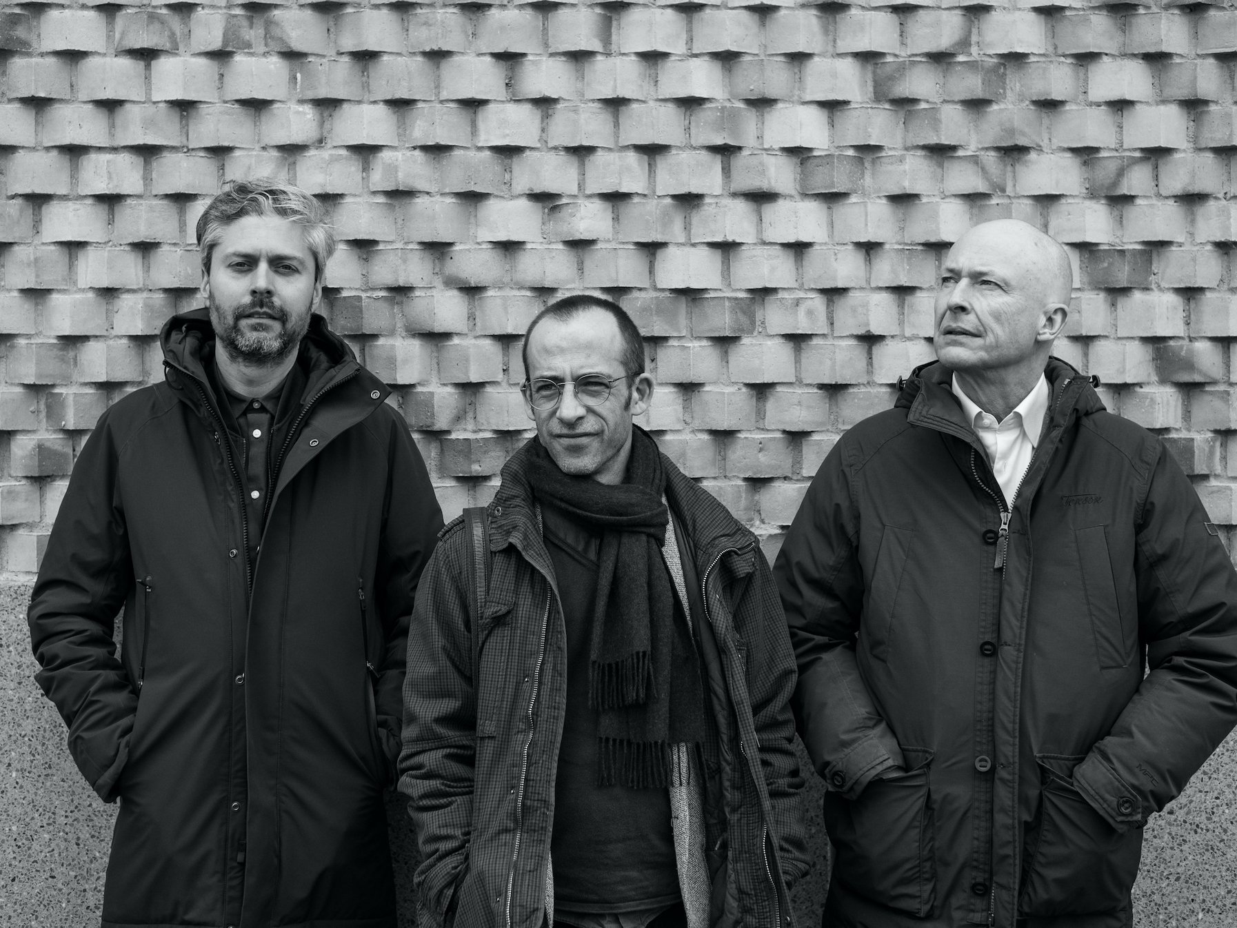 Three men standing outdoors against a brick wall, wearing winter jackets.