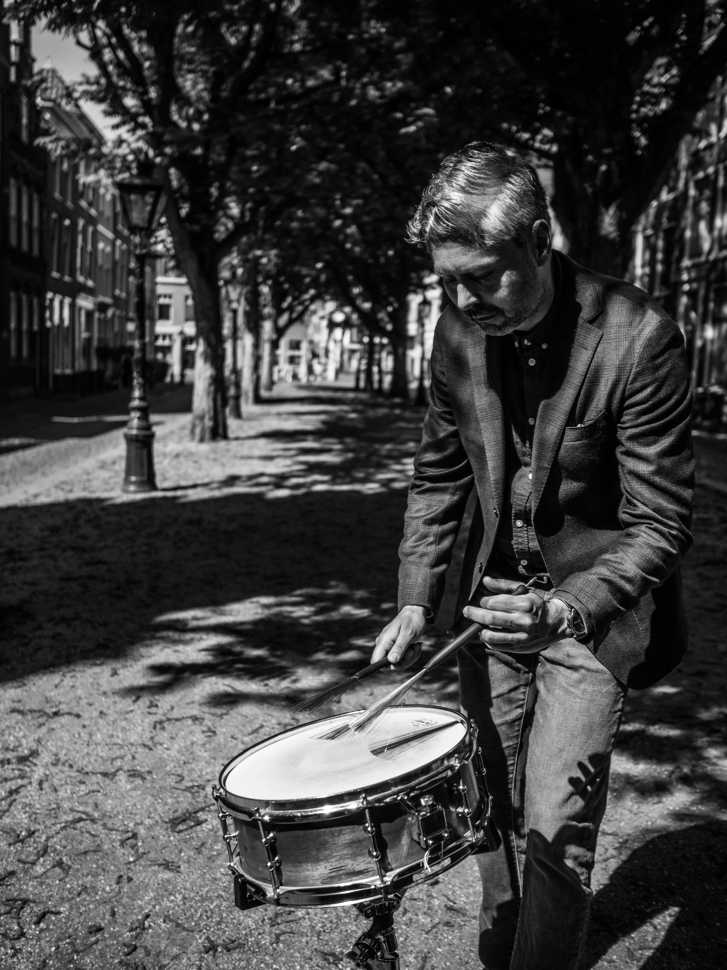 A man in a suit playing a drum on a city street lined with trees.