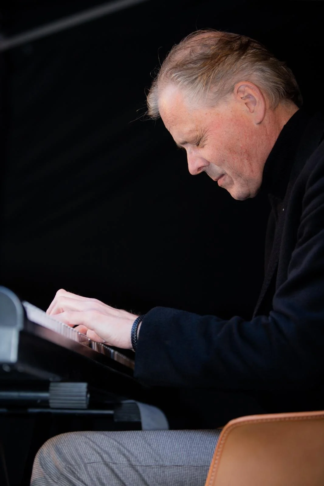 A man playing the piano, focused on playing keyboard, wearing a black coat, sitting on a beige chair.