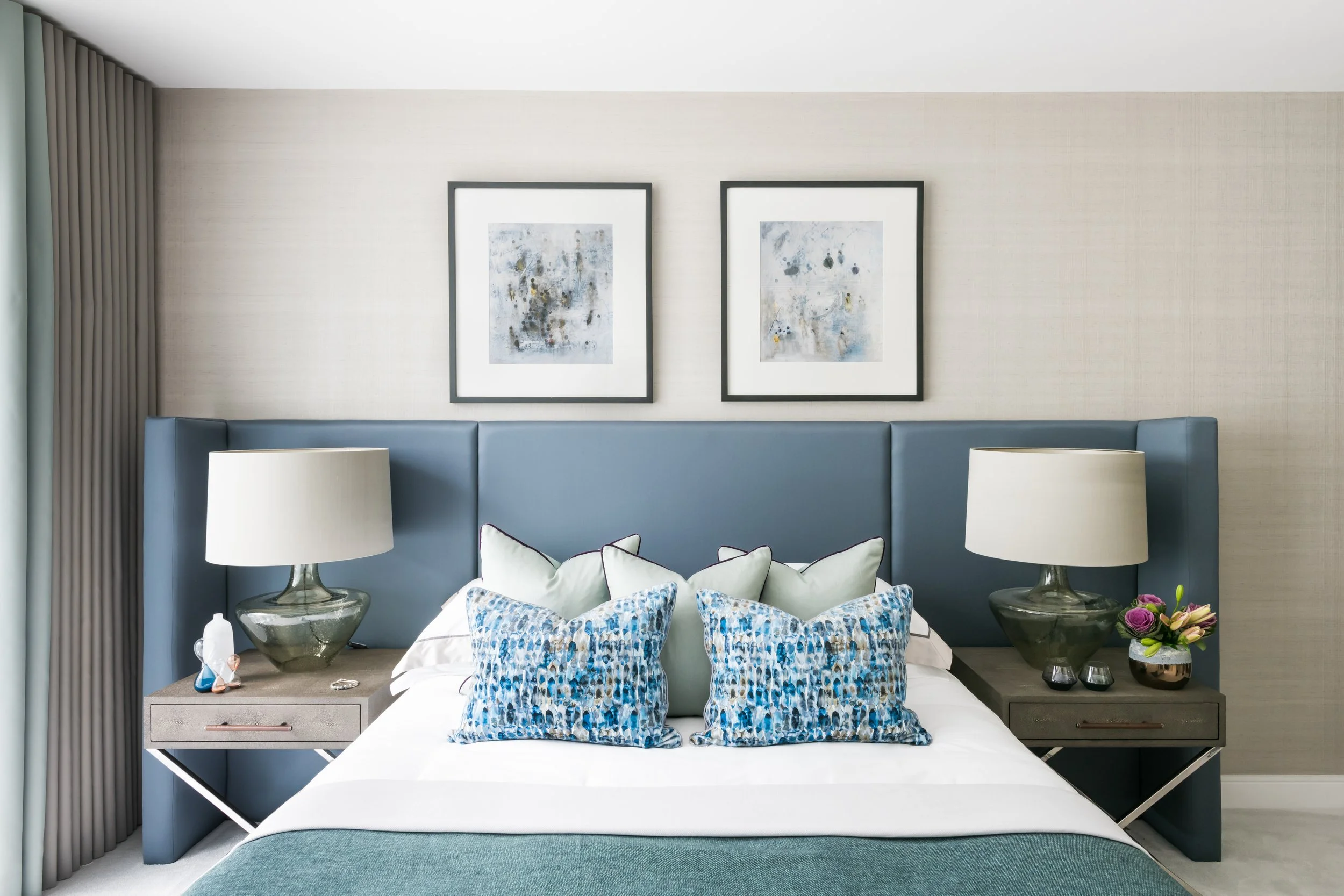 Modern bedroom with a blue upholstered headboard, twin nightstands with lamps, and abstract artwork above the headboard. The bed has decorative pillows with blue patterns, and there is a small vase with flowers on one of the nightstands.