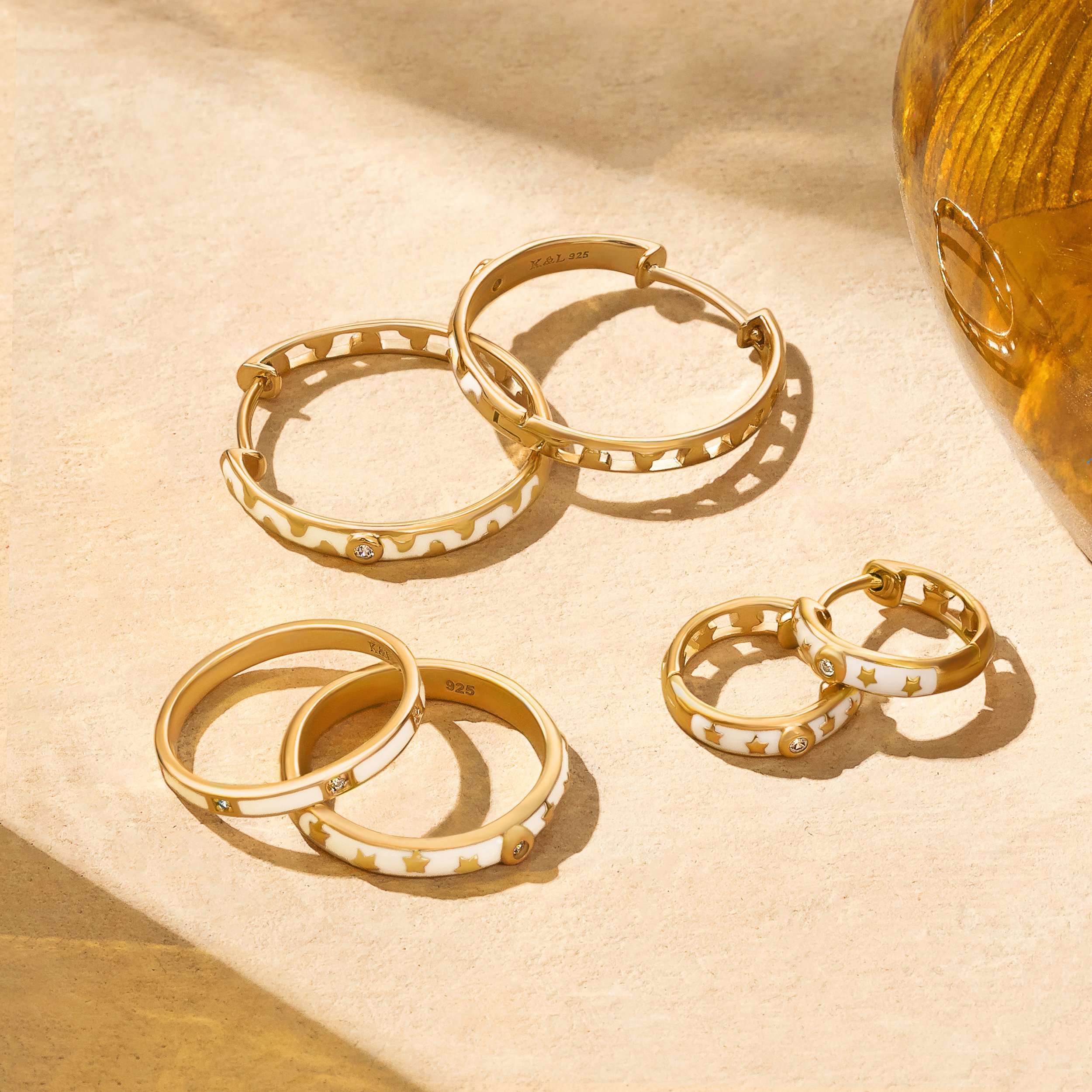 Gold hoop earrings with star and dot designs and embedded rhinestones, placed on a beige surface with a amber-colored glass container nearby.