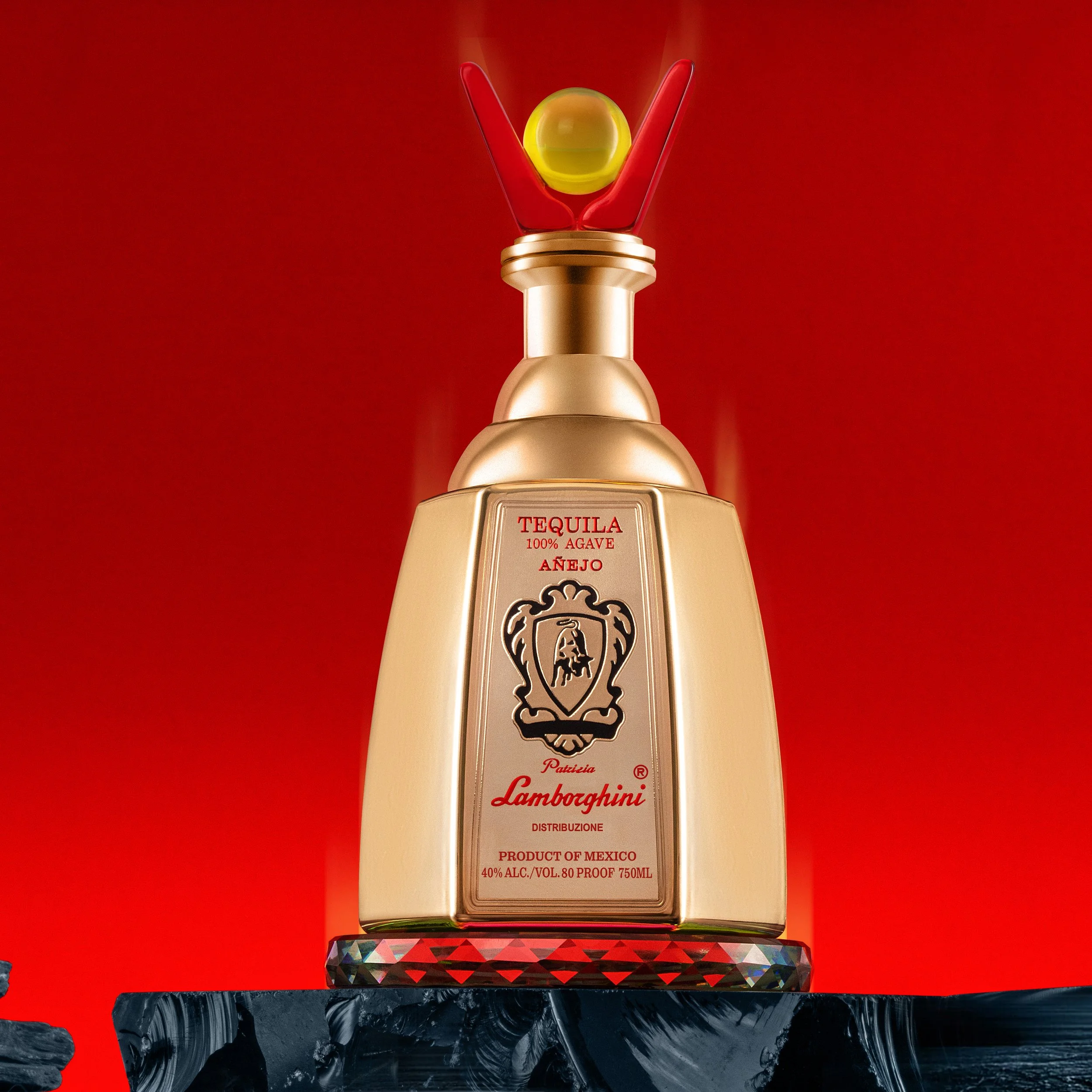 A large decorative bottle with a gold finish, resembling a liquor bottle, featuring a label that reads "Tequila 100% Agave Añejo" with a Lamborghini logo and text indicating it is a product of Mexico with 40% alcohol content. The bottle has a yellow and red ball on top, surrounded by a red stick, set against a red background, and is placed on a decorative black and multicolored stand.