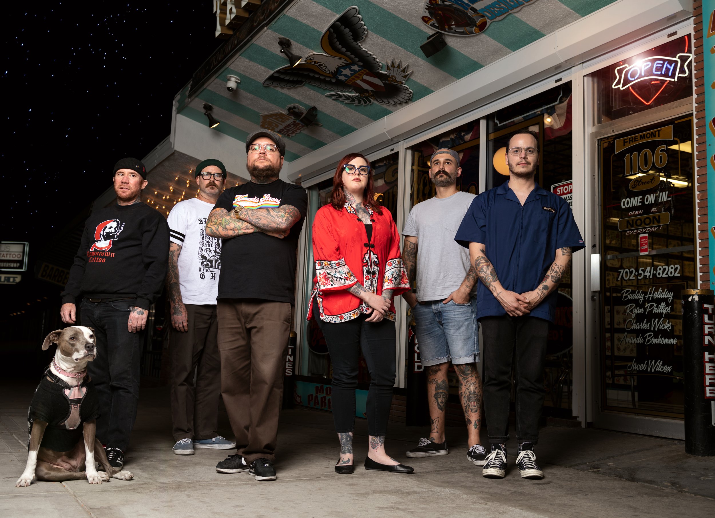 Group of six people standing in front of a vintage shop at night, with small dog sitting on the sidewalk. The people include five men and one woman, dressed casually, some with tattoos, standing with arms crossed or hands in pockets, and a dog wearing a harness. The shop has an open neon sign and colorful signage, with outdoor decorative elements, and a starry night sky is visible above.