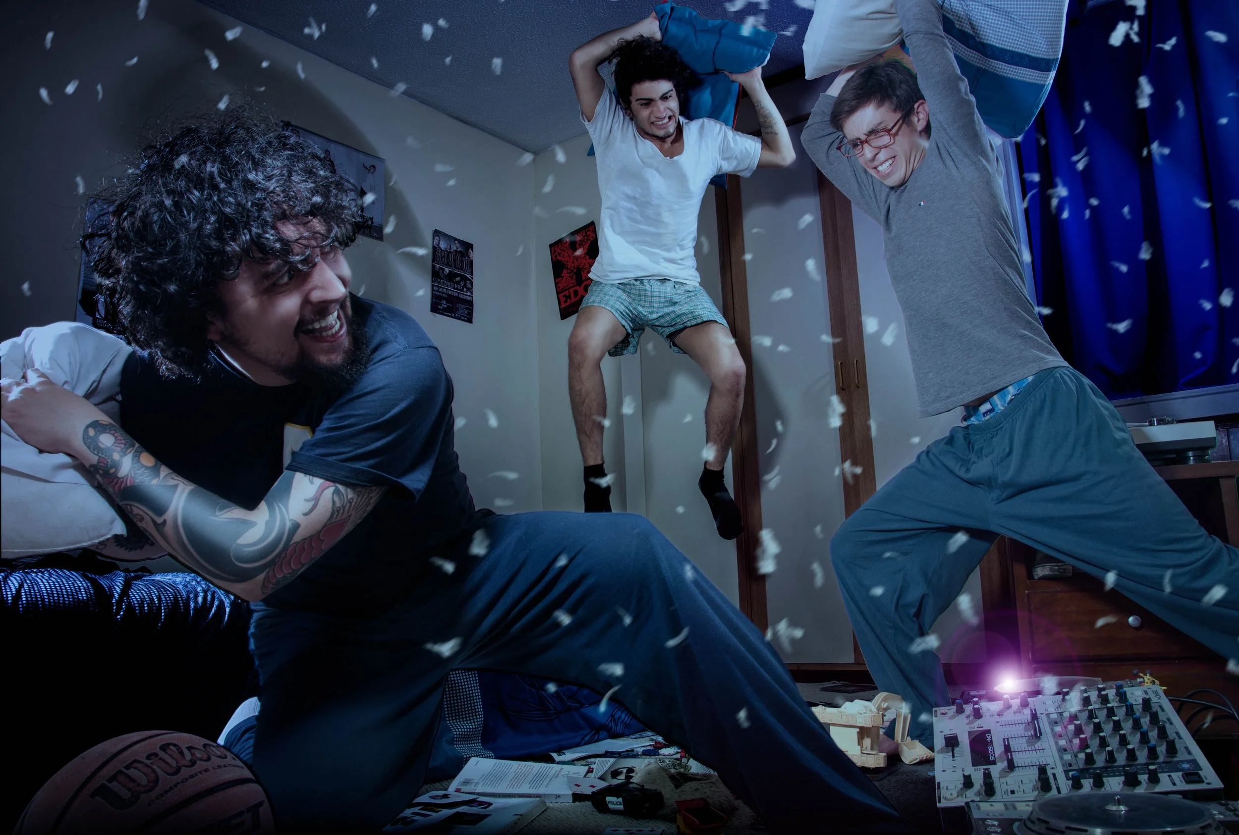 Three young men having a lively indoor party with pillows and confetti, one standing on a bed, two dancing in front of a DJ mixer, in a room with posters on the wall and a basketball on the floor.