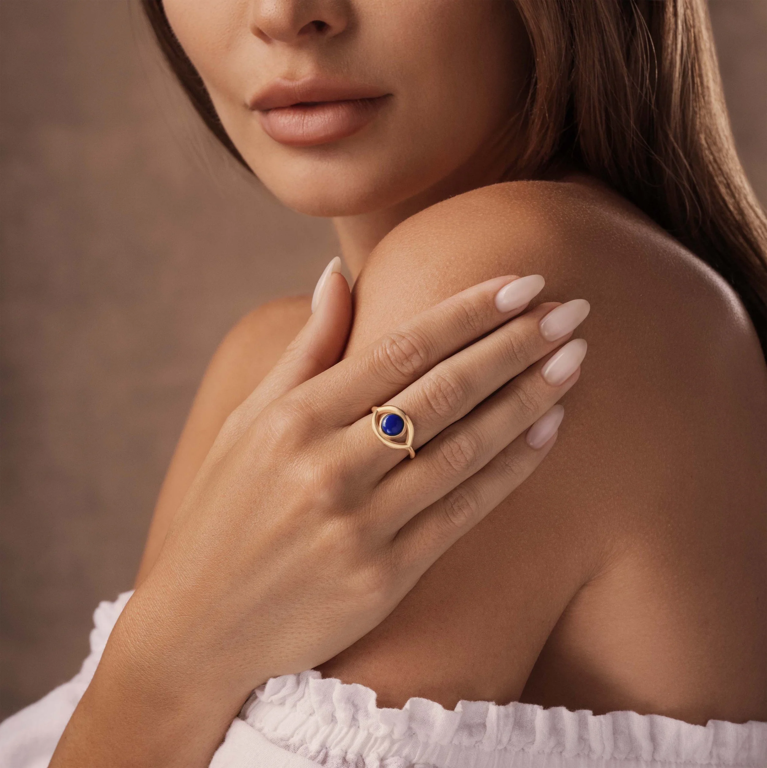 A close-up of a woman with fair skin, holding her shoulder with her hand. She is wearing a blue ring with a gold band and has neatly manicured nails. She has brown hair and is wearing a white off-the-shoulder top.