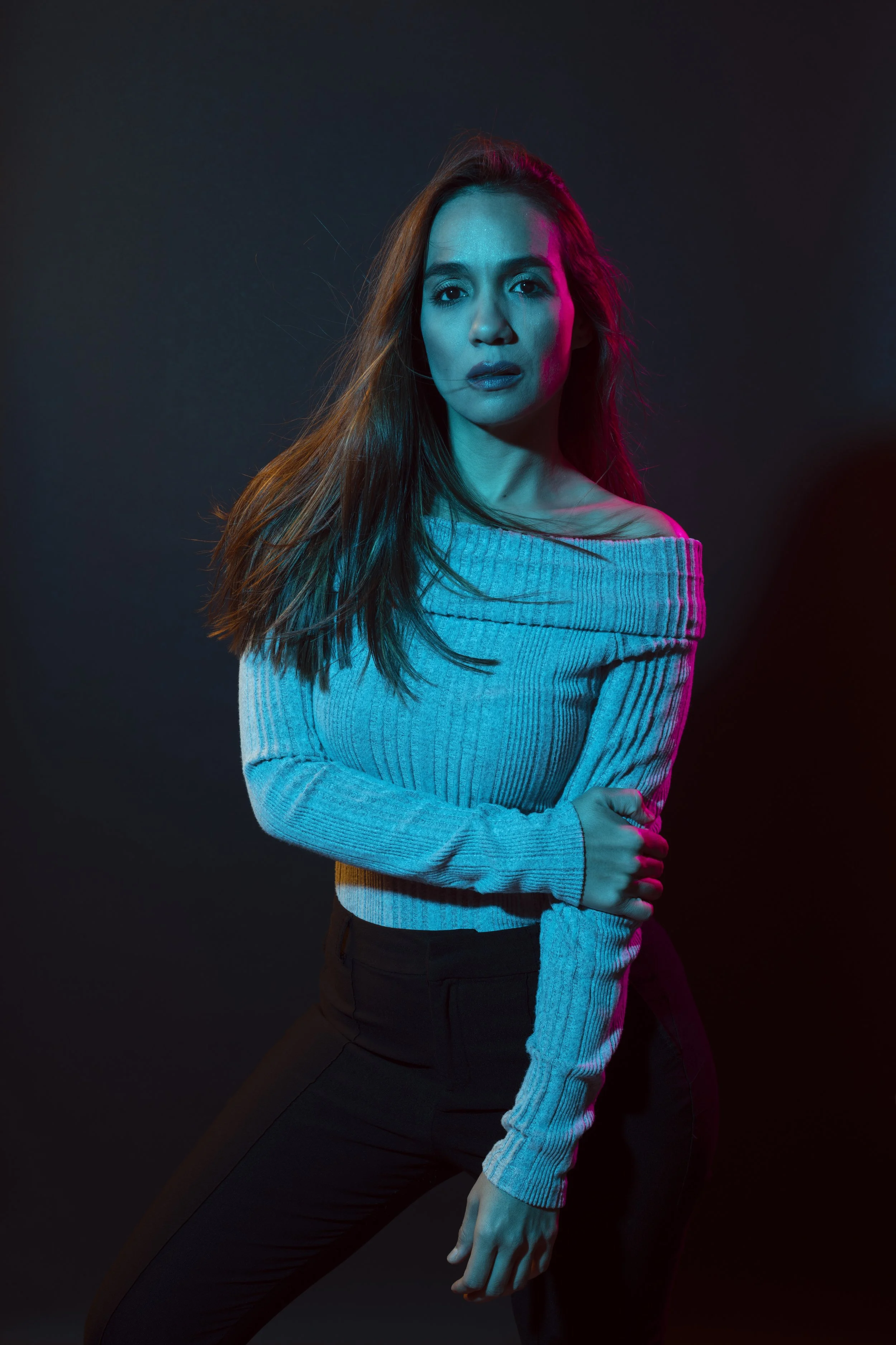 A woman with long hair in a gray sweater and black pants posing against a dark background with colorful lighting.
