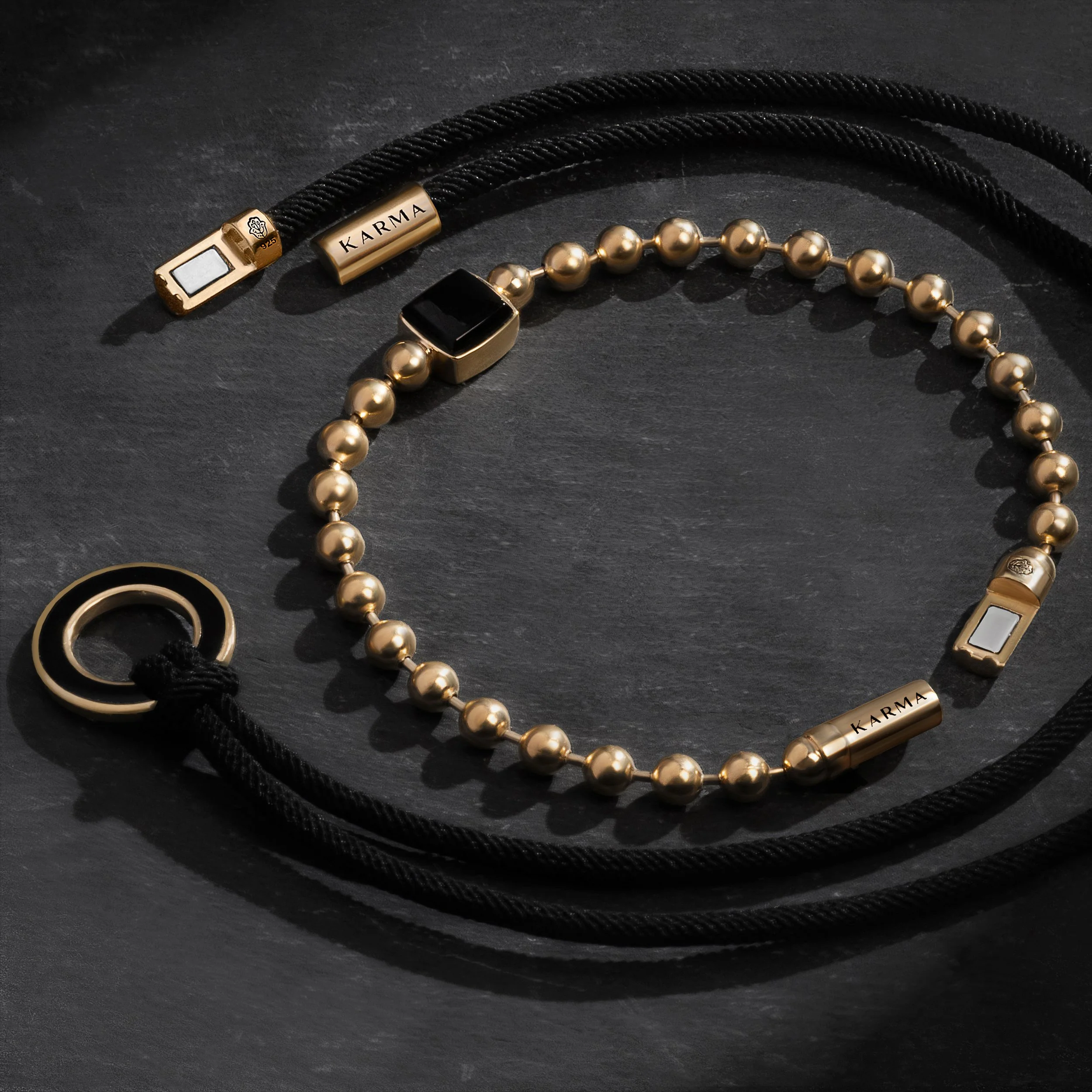Set of men's jewelry including a black beaded necklace with gold accents, a black beaded bracelet, and a gold ring with a black stone, all on a black surface.