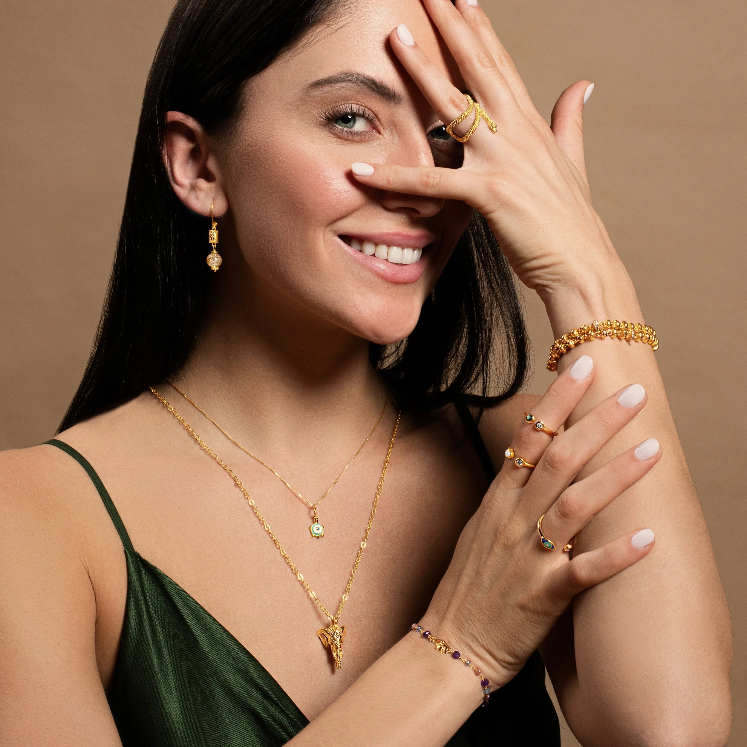 A woman with dark hair wearing multiple gold jewelry pieces, including necklaces, rings, earrings, bracelets, and an ornate gold ring on her index finger, is smiling and posing with her hand covering part of her face against a beige background.