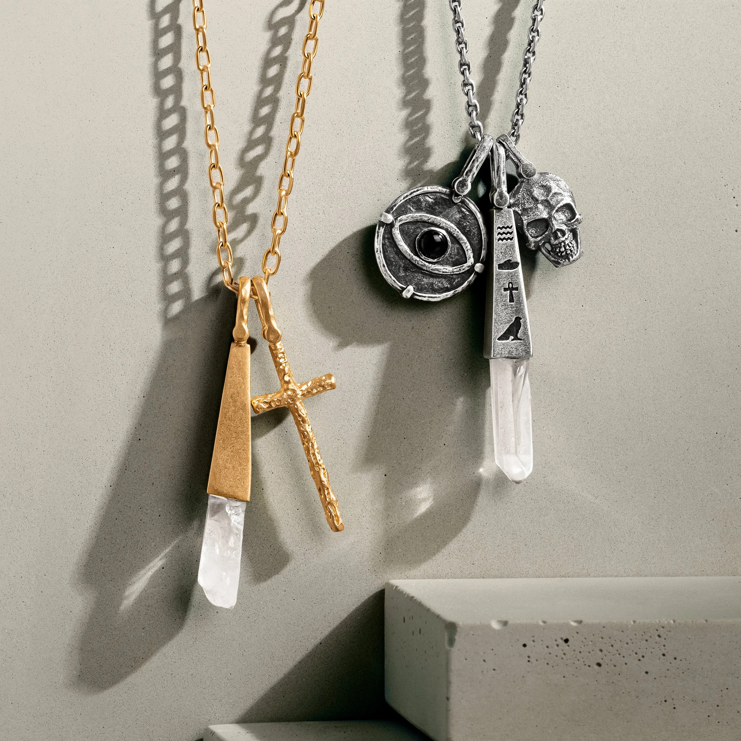 Two necklaces hanging on a plain wall with shadows cast by the jewelry. The left necklace is gold with a cross and crystal pendant, and the right necklace is silver with an eye, skull, and crystal pendant.