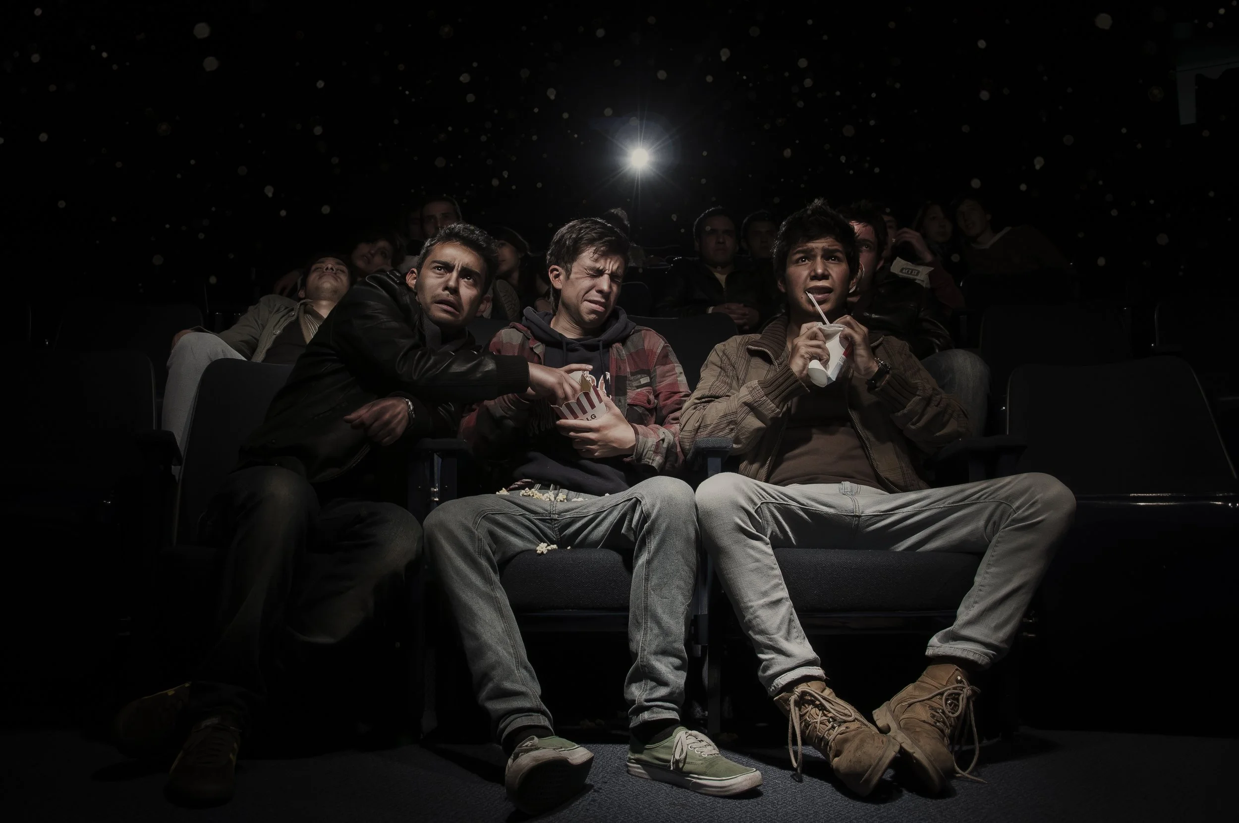 Three young men sitting in a dark movie theater, watching a film, with popcorn and drinks, reacting with surprise or concern.