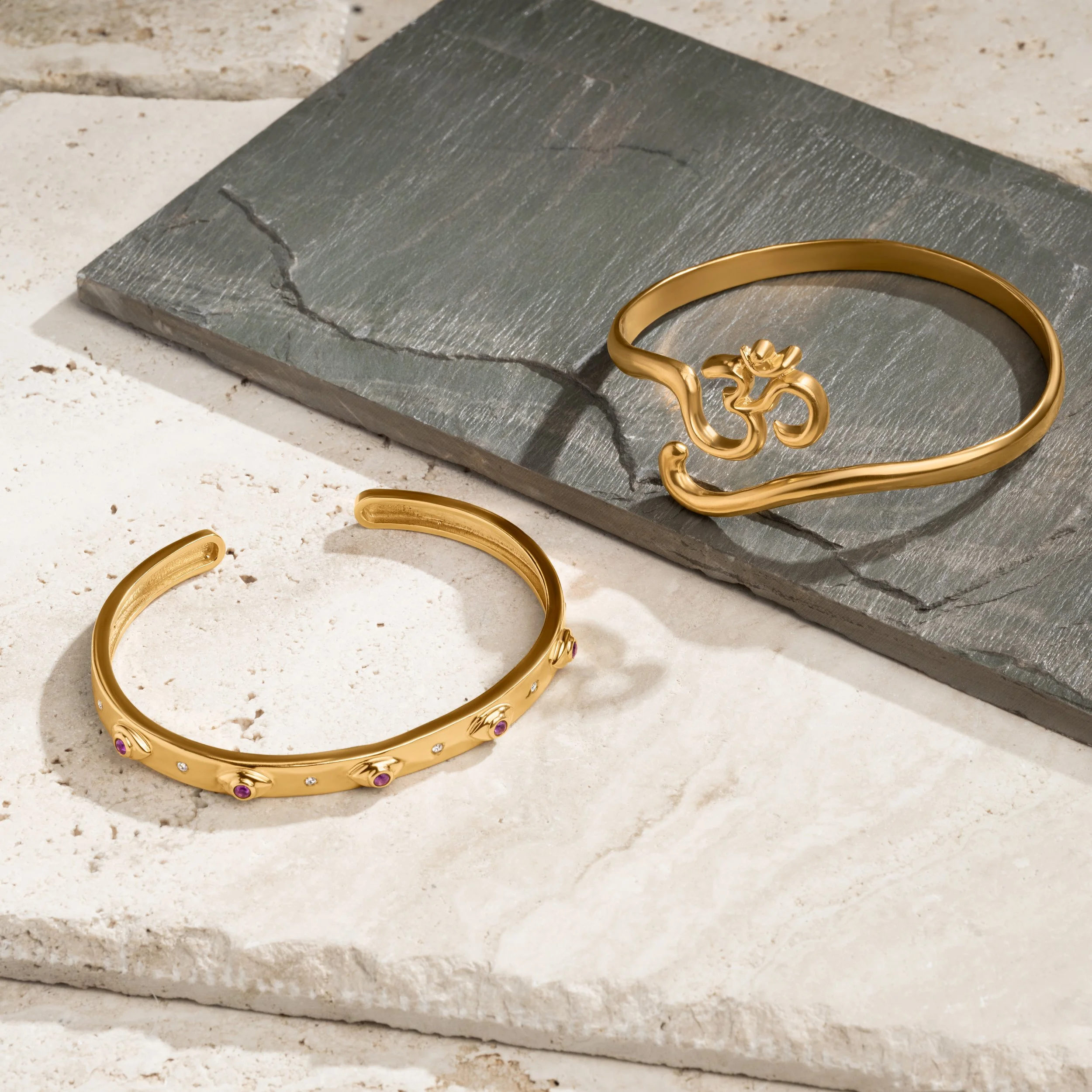 Gold jewelry pieces, including a bracelet with purple gemstones and a cuff with an Om symbol, displayed on stone and concrete surfaces.