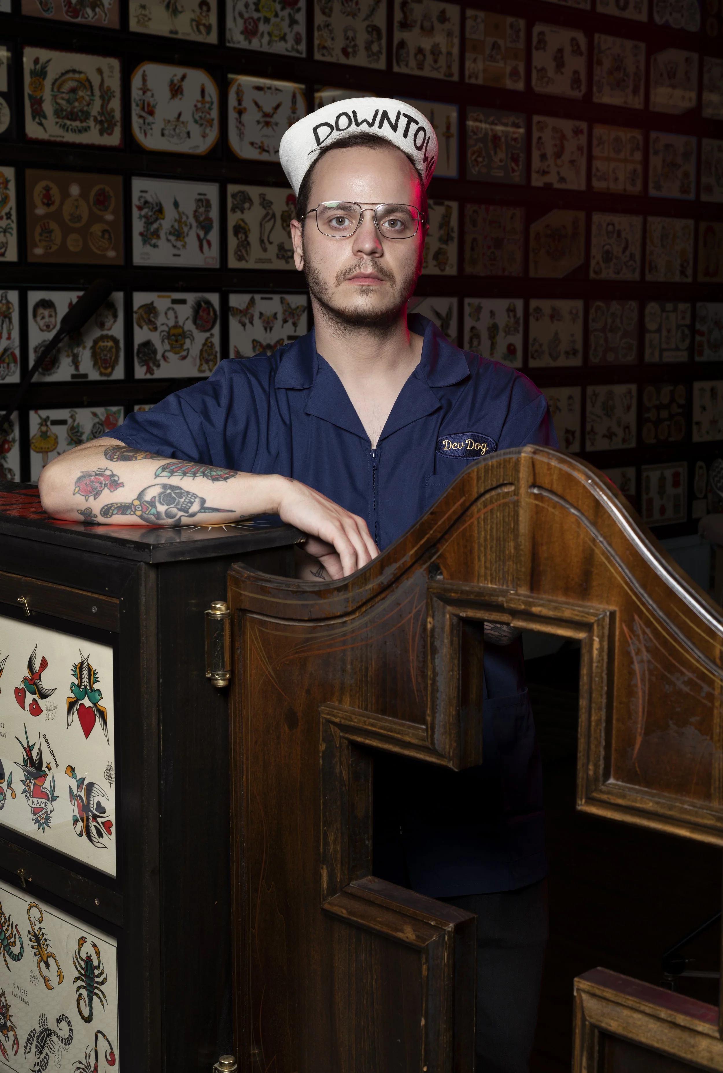 A man with glasses and tattoos, wearing a navy blue shirt and a white hat with 'DOWNTOWN' written on it, leaning on a tattoo-style decorated counter in a tattoo shop with tattoo flash art on the walls.