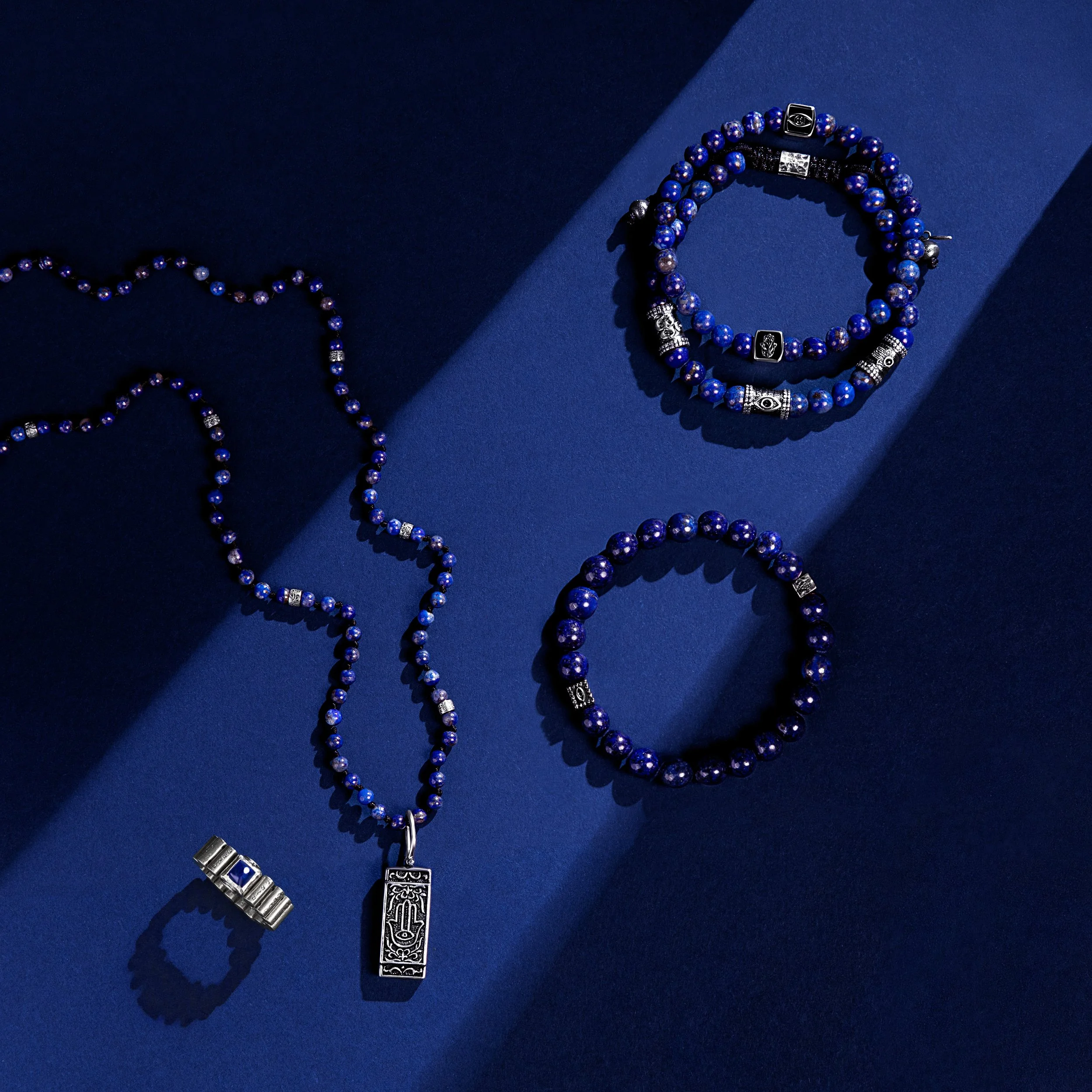 Blue beaded jewelry set including a necklace, two bracelets, a ring, and a pendant with silver accents, all arranged on dark blue background.