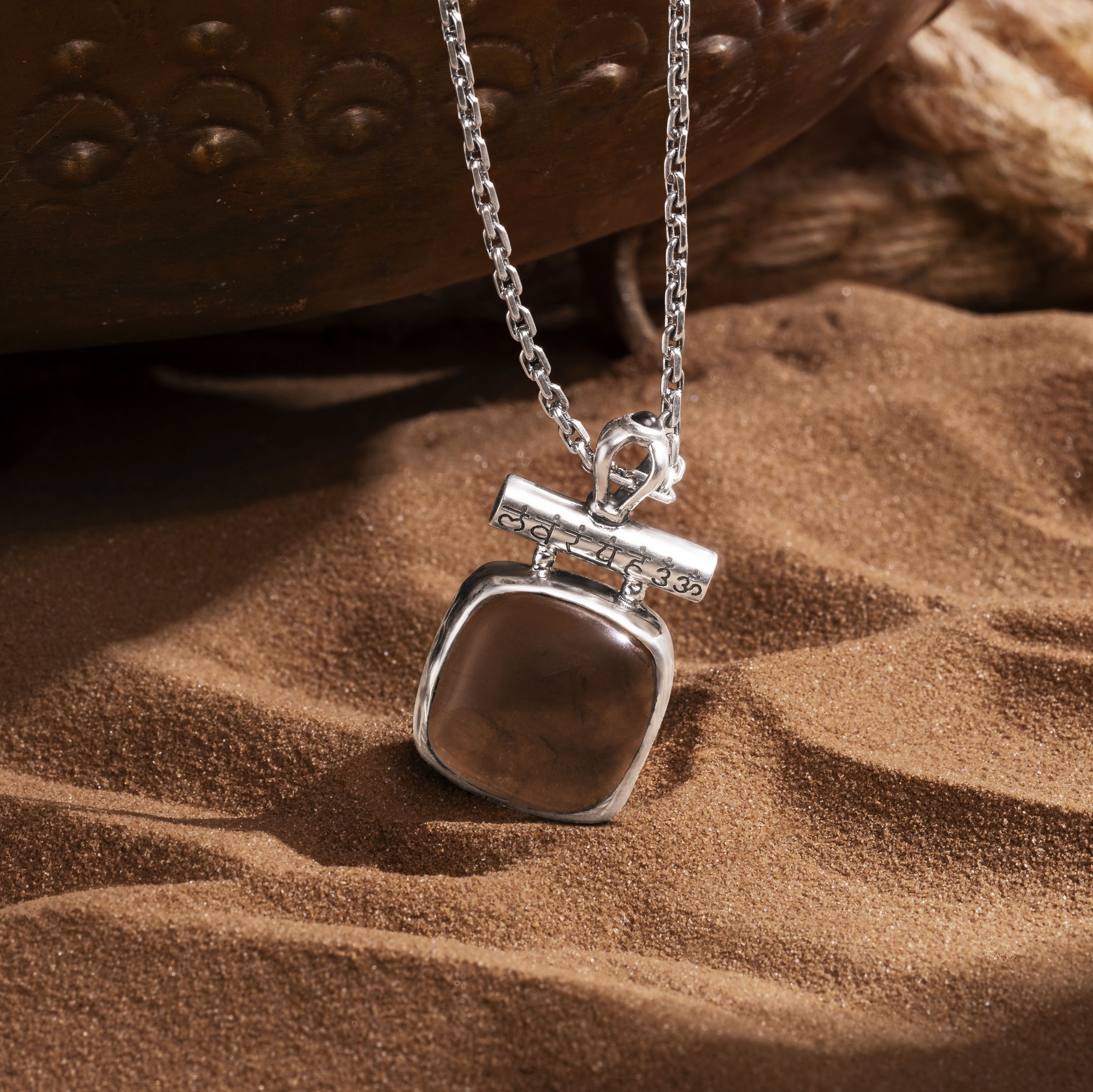 A silver necklace with a square brown pendant, resting on a sandy surface with a textured wooden object in the background.