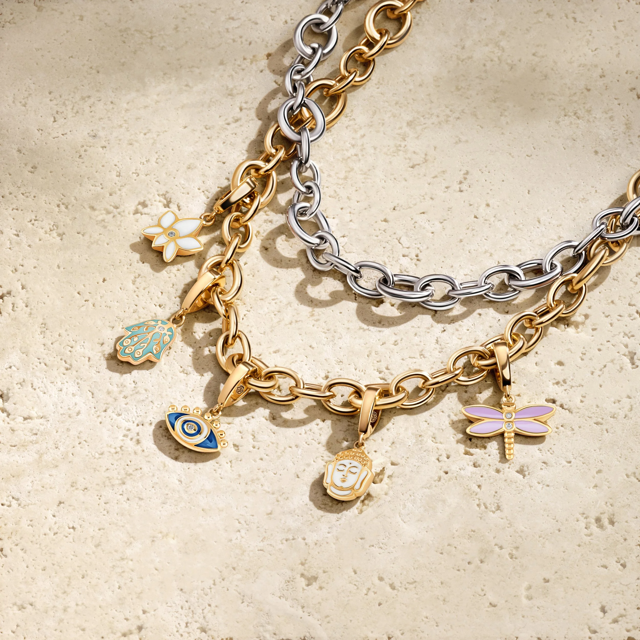 Three layered metal chain necklaces with various charms on a sandy surface, featuring gold and silver tones, and charms including a flower, leaf, eye, face, and a dragonfly.