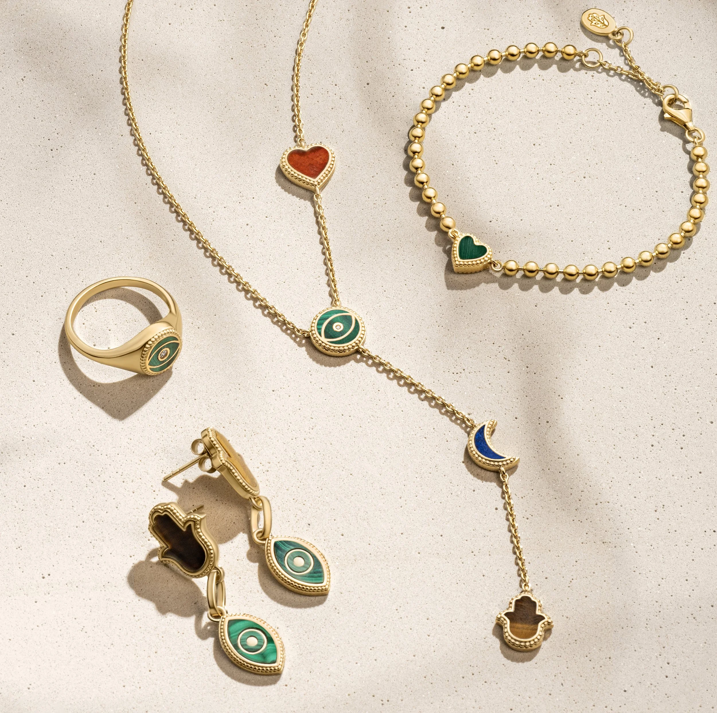 Gold jewelry set including a ring, earrings, necklace, bracelet, and a chain with colorful heart and moon pendants and eye motifs, laid out on a light-colored surface.