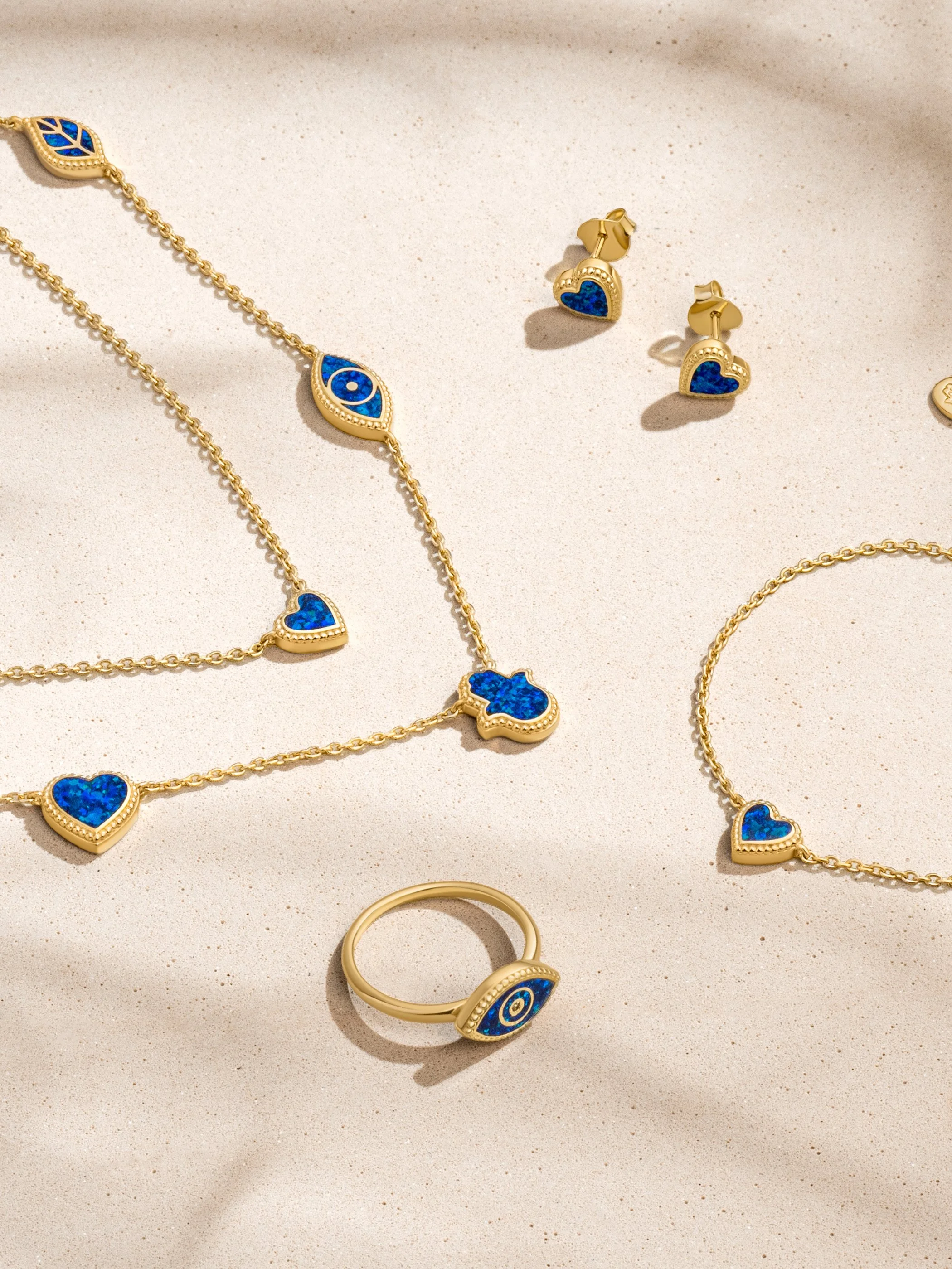 Gold jewelry set including a necklace, earrings, bracelet, and ring, all featuring blue heart and eye-shaped charms.