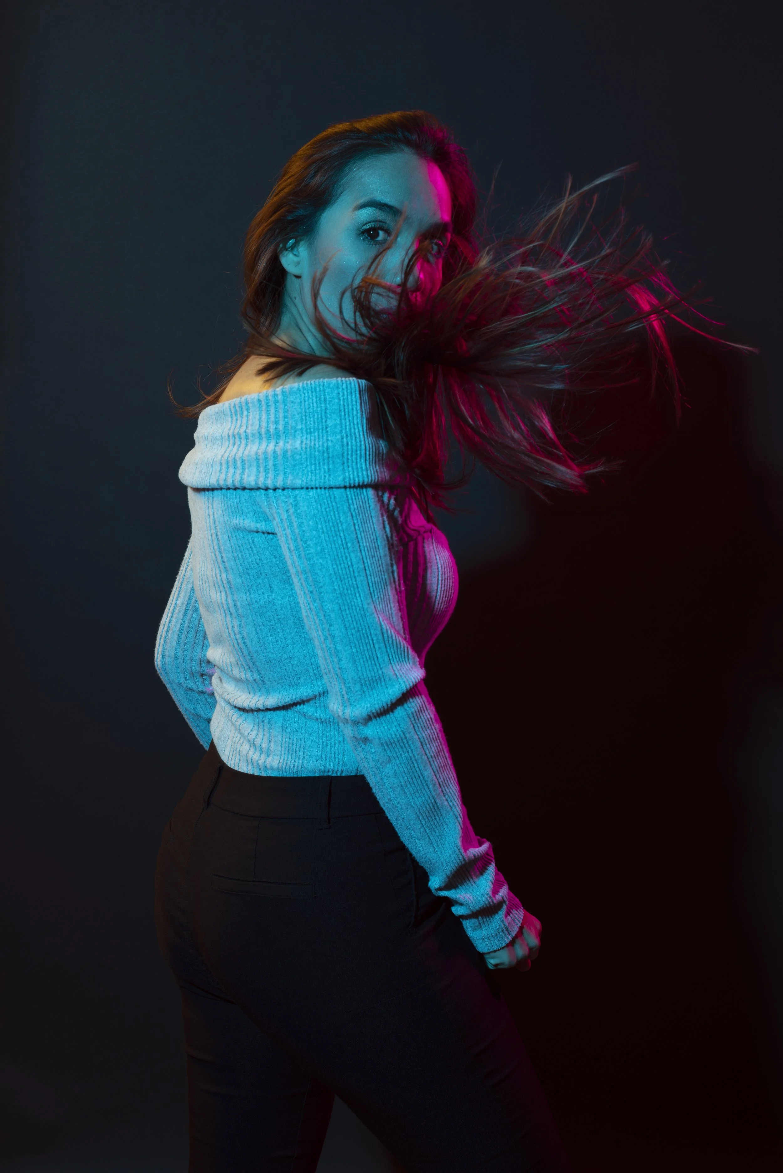 A woman with long brown hair, wearing a light blue off-the-shoulder sweater and black pants, is looking over her shoulder as her hair is thrown in motion, illuminated by pink and blue lights against a dark background.