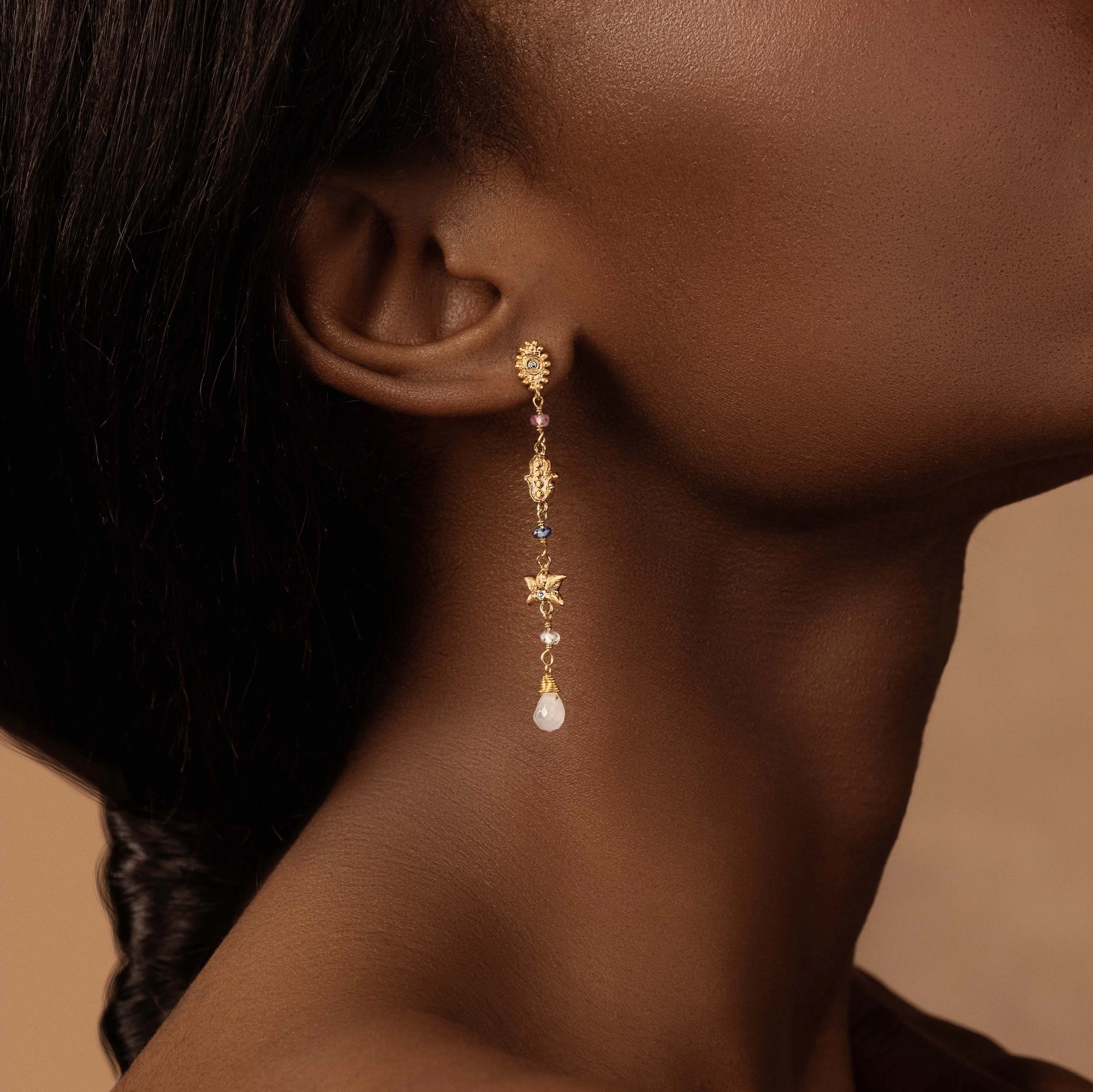 Close-up of a woman's ear wearing a long, dangling, gold earring with various colorful charms and a teardrop-shaped pearl at the end.
